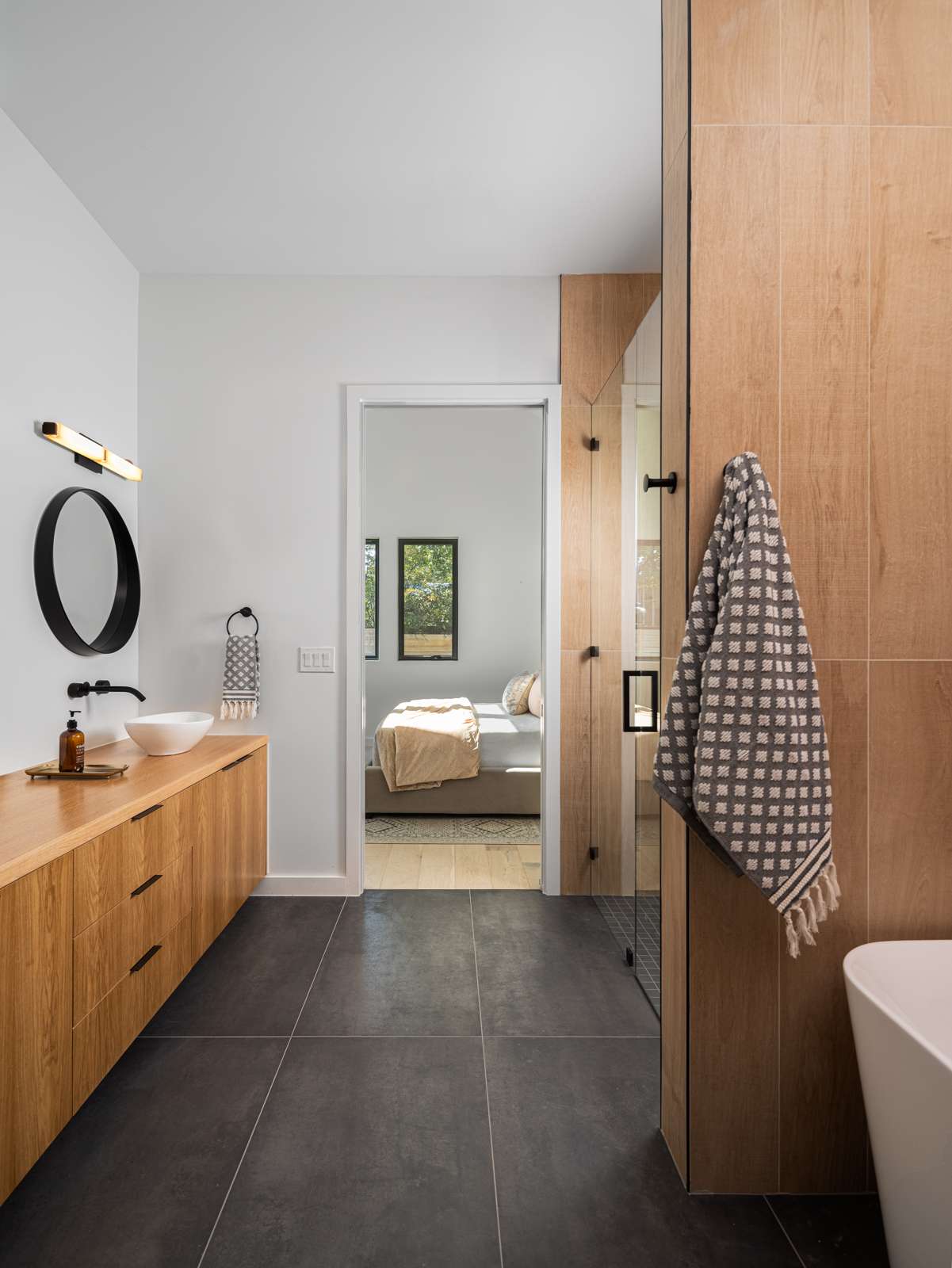 Minimalist ensuite bathroom with warm wood cabinetry, sleek black fixtures, and oversized charcoal floor tiles for a clean, modern look. A vessel sink and round mirror add sculptural elegance, while the walk-in shower features natural wood-look tile for a spa-like vibe. The open doorway leads seamlessly into a cozy, light-filled bedroom, creating a connected and calming primary suite.