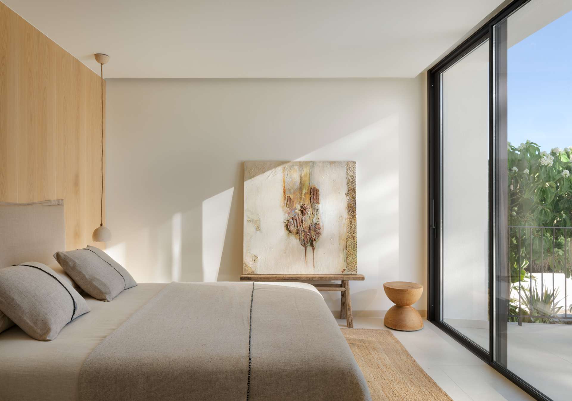 The design of this bedroom is an exercise in restful restraint. Sunlight filters through sliding doors that open onto a balcony surrounded by lush greenery, while soft linen bedding, natural textures, and minimal styling keep the mood grounded. A timber feature wall adds quiet warmth, and sculptural accents, from side tables to abstract art, offer subtle personality within the calm palette. It’s a room designed for retreat, where every element supports peace.