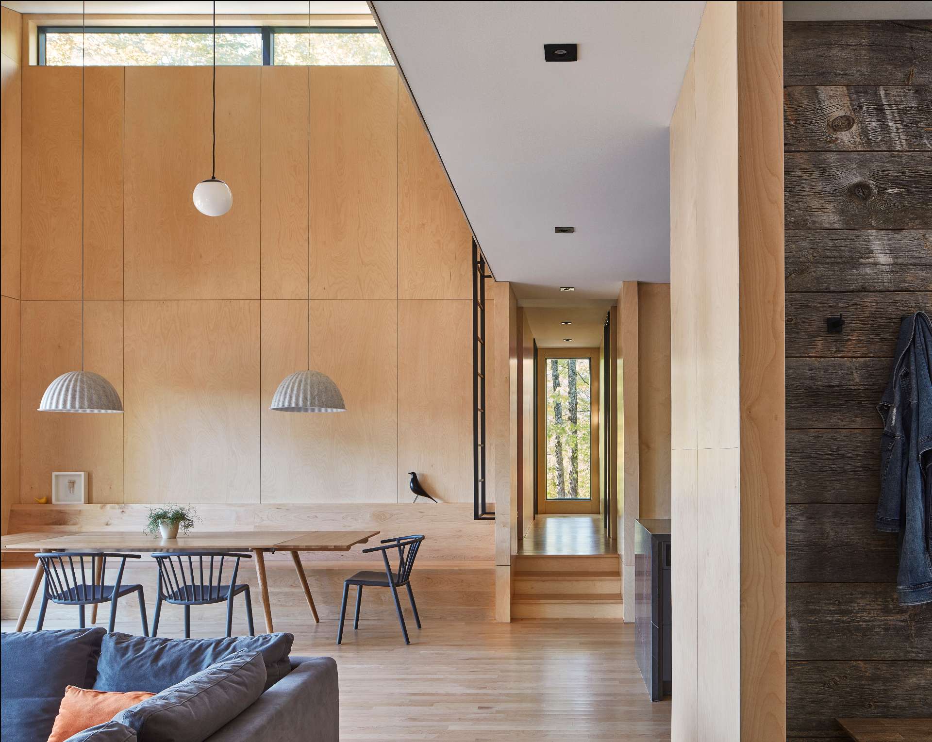 At the center of the living space in this modern cottage, the dining area includes a built-in maple plywood banquette, creating a warm and inviting atmosphere for gatherings around the dining table.
