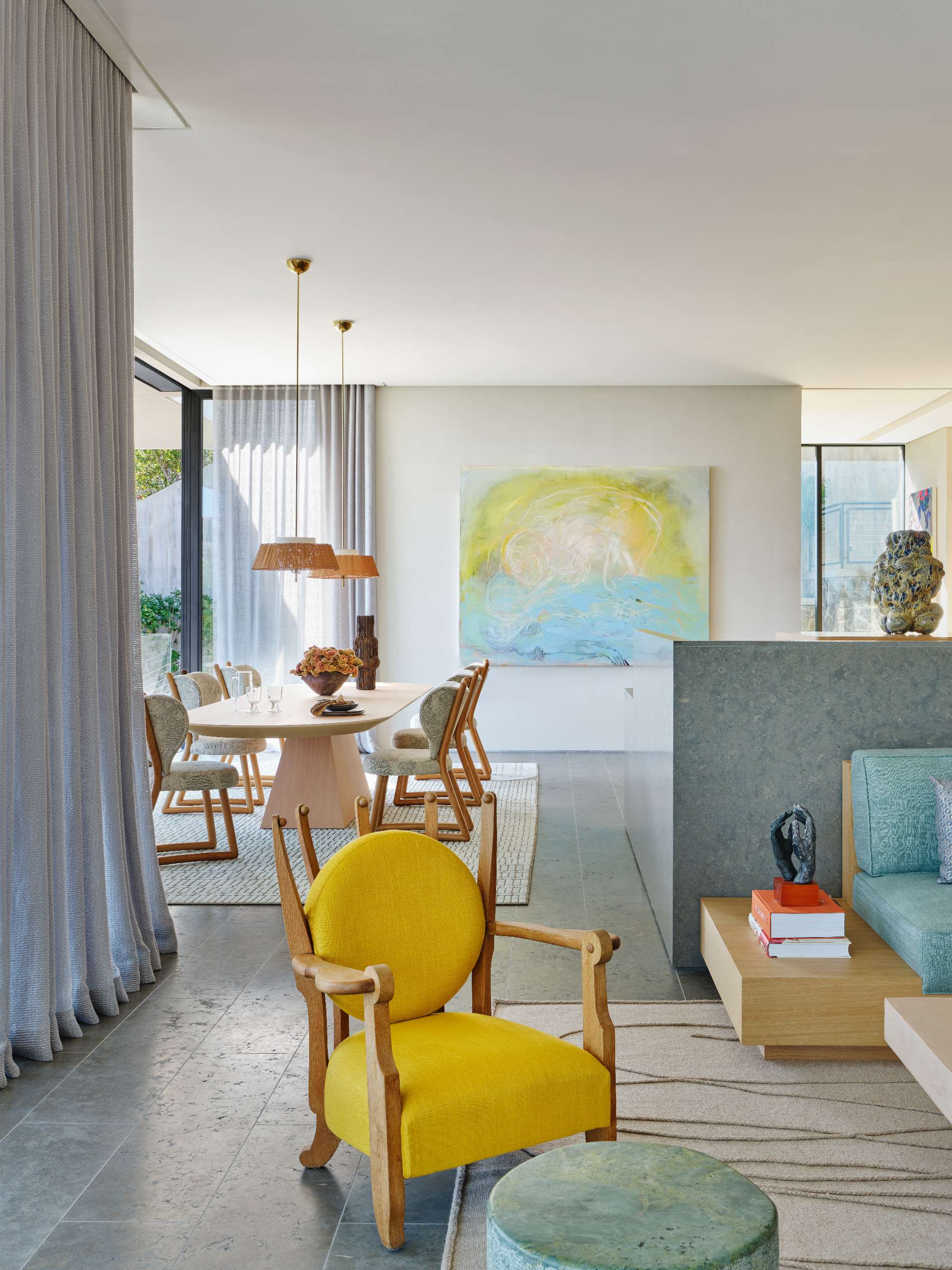 The dining room combines soft textures and natural materials for a relaxed, coastal feel. A sculptural wood table anchors the space, surrounded by upholstered chairs with warm wood frames. Sheer drapery filters the light, while woven pendant lights and a colorful abstract painting add layers of warmth and character.