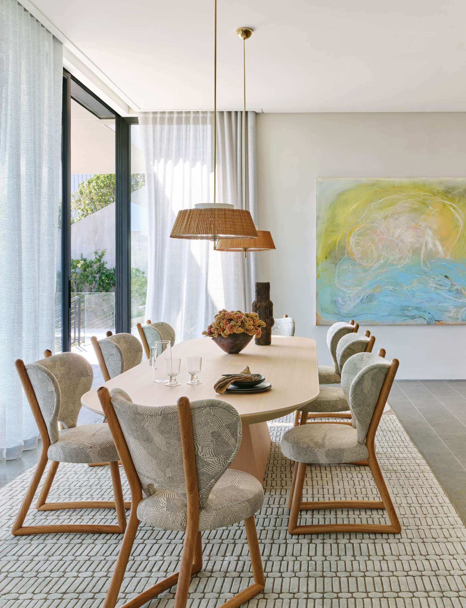The dining room combines soft textures and natural materials for a relaxed, coastal feel. A sculptural wood table anchors the space, surrounded by upholstered chairs with warm wood frames. Sheer drapery filters the light, while woven pendant lights and a colorful abstract painting add layers of warmth and character.