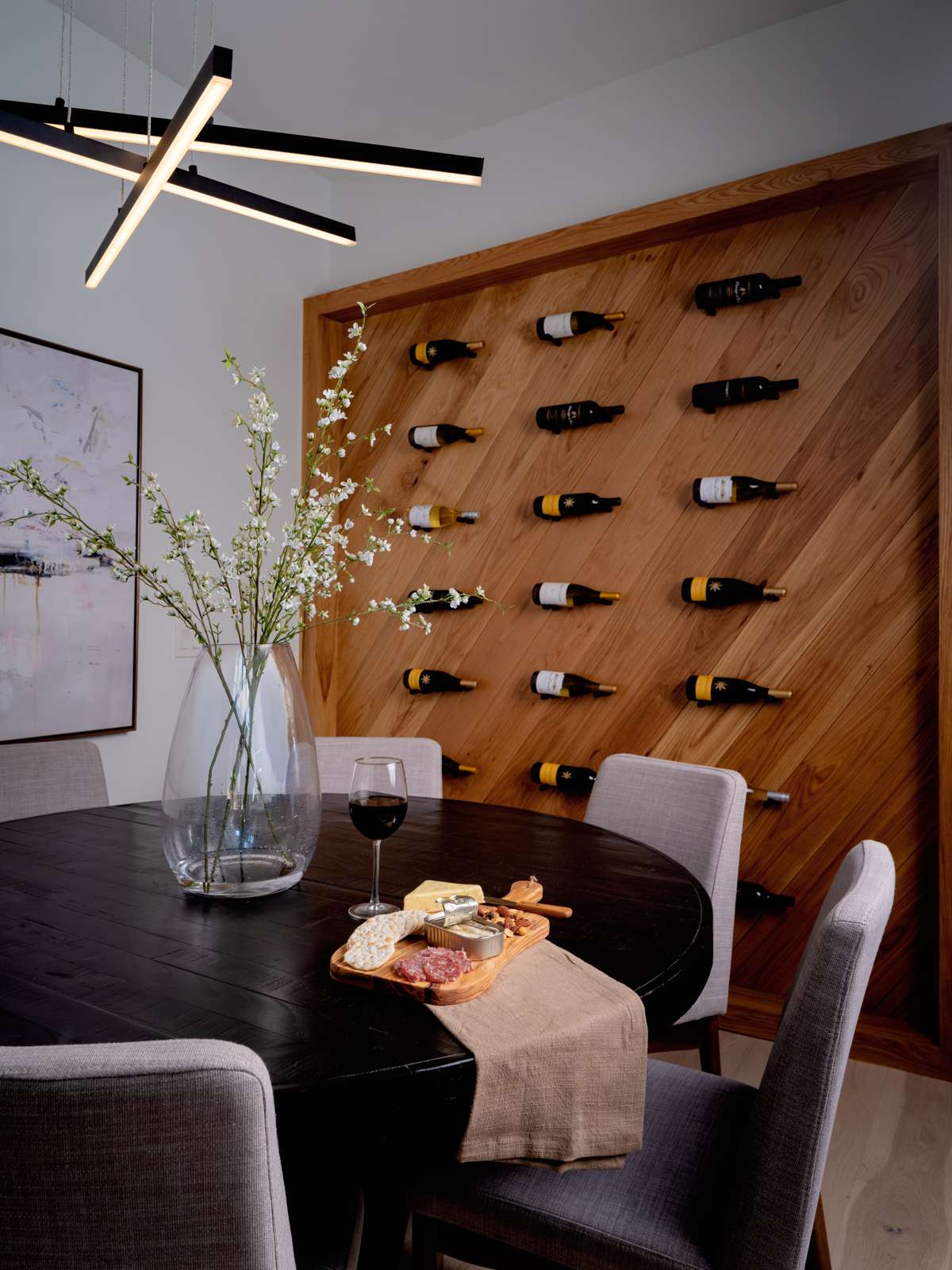 This dining room pairs modern simplicity with warmth and texture. A built-in diagonal wood wine wall creates a striking focal point and adds rich texture. A round dark table is surrounded by soft gray chairs, while overhead, a sculptural pendant light brings a contemporary finish to the space, designed with gathering and everyday elegance in mind.