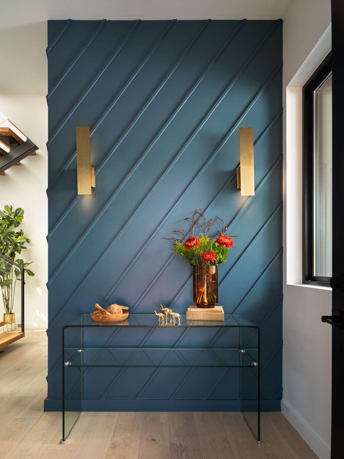 The entryway sets the tone with a bold blue accent wall detailed in diagonal paneling. Sleek brass sconces bring a soft glow, while a glass console table keeps the space feeling open and light. Sculptural objects and fresh florals add a thoughtful touch, creating an inviting first impression in a compact footprint.
