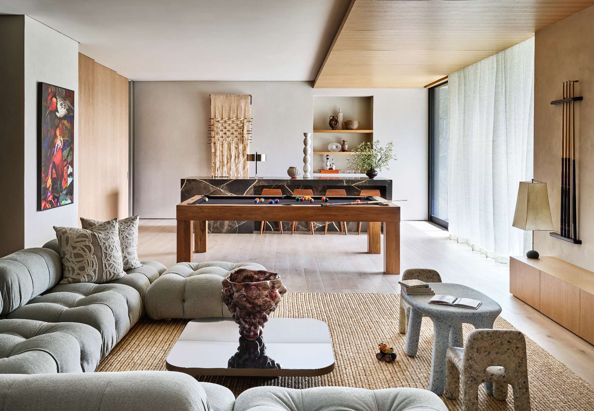 A home bar anchors the far end of the open-plan living space, while open shelving niche showcases a curated display of ceramic vessels and glassware. A streamlined wooden pool table introduces a playful touch, complemented by a plush sofa and layered textures that lend the room a relaxed yet refined feel.