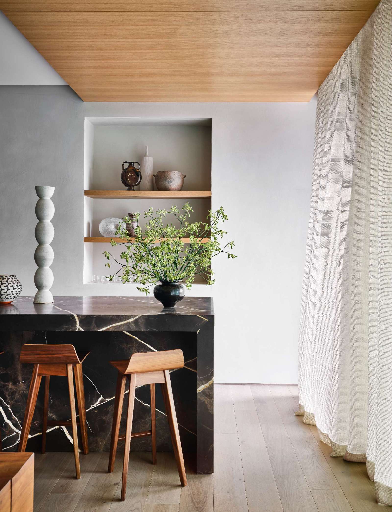 A home bar anchors the far end of the open-plan living space, while open shelving niche showcases a curated display of ceramic vessels and glassware.