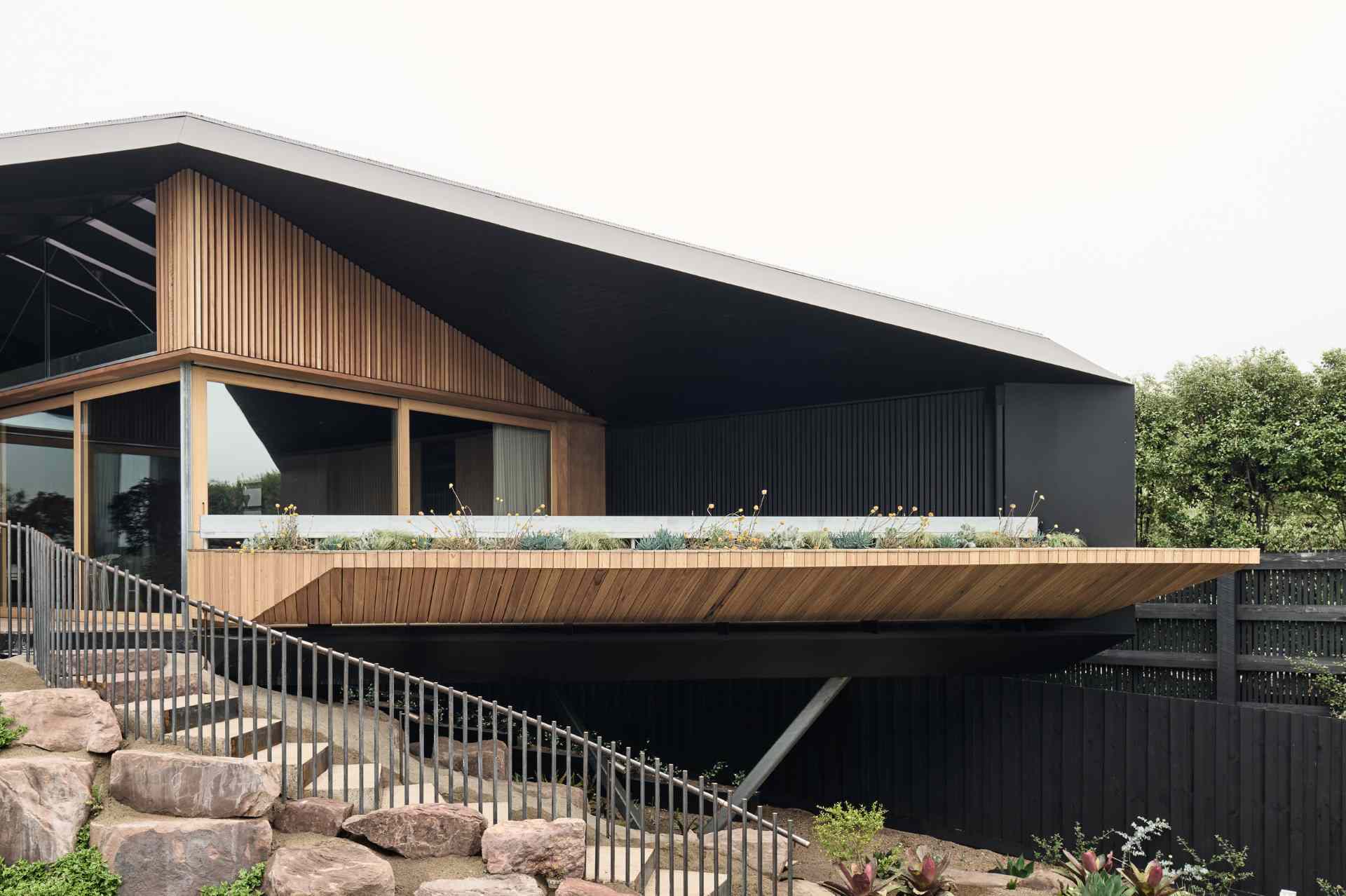 The rear facade blends bold architecture with the natural landscape. An angular roof hovers over decks and planters, while a curving stairway winds through native plantings. Below, a sleek lap pool runs along the slope, grounding the structure in its coastal setting.