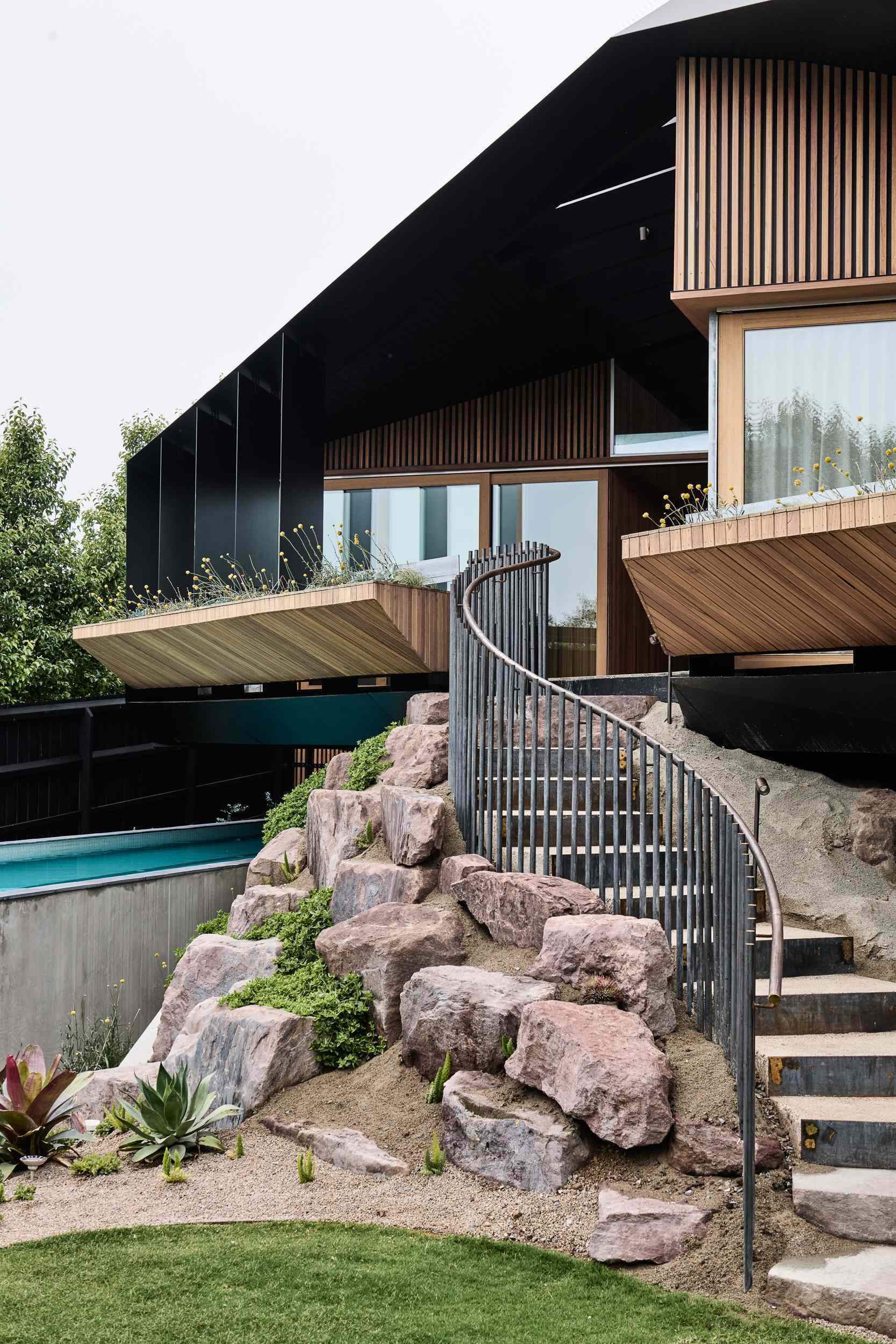 The rear facade blends bold architecture with the natural landscape. An angular roof hovers over decks and planters, while a curving stairway winds through native plantings. Below, a sleek lap pool runs along the slope, grounding the structure in its coastal setting.