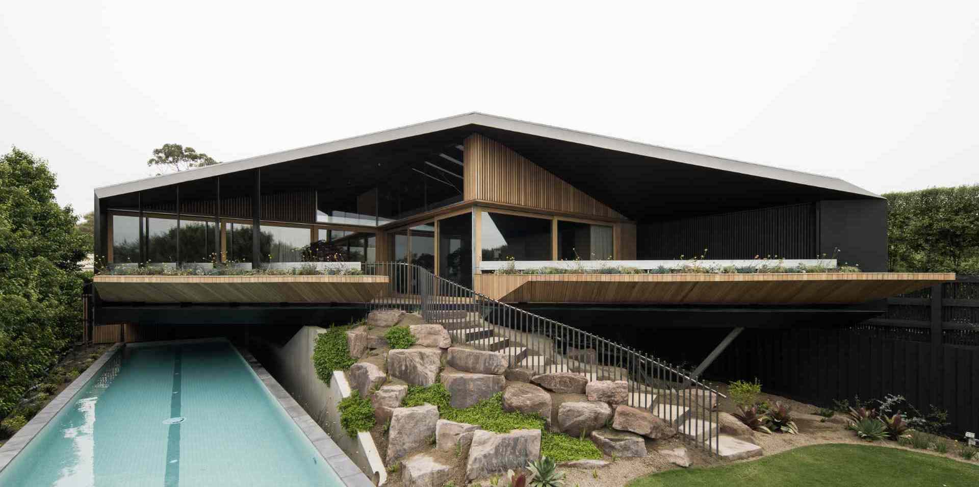 The rear facade blends bold architecture with the natural landscape. An angular roof hovers over decks and planters, while a curving stairway winds through native plantings. Below, a sleek lap pool runs along the slope, grounding the structure in its coastal setting.
Below, a lap pool runs parallel to the structure, tucked elegantly into the base of the slope. Together, the layered architecture, natural stonework, and linear pool create a composition that feels both grounded and elevated.