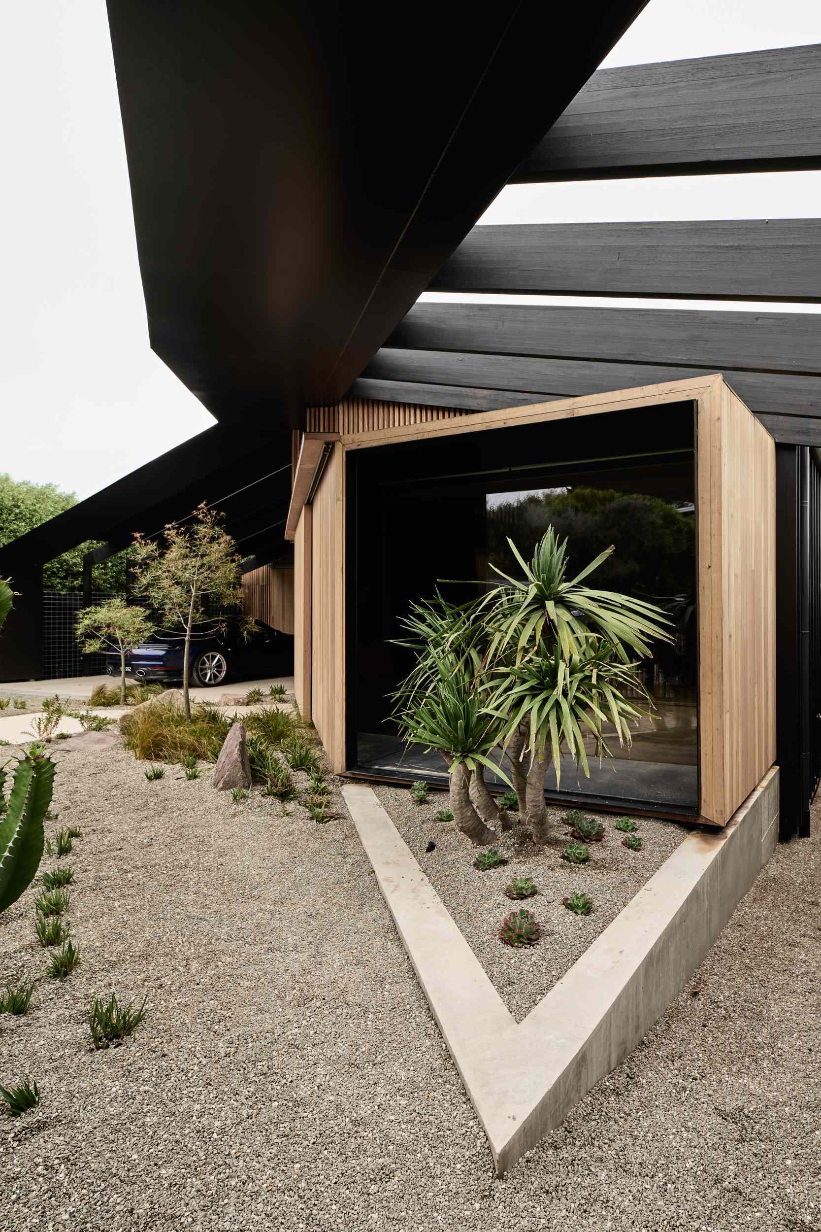 A striking picture window extends from the facade, framing serene views and filling the interior with natural light. Below, native landscaping with gravel and exposed rocks enhances the coastal feel, while a sculptural concrete planter near the entry mirrors the home’s crisp, geometric design.