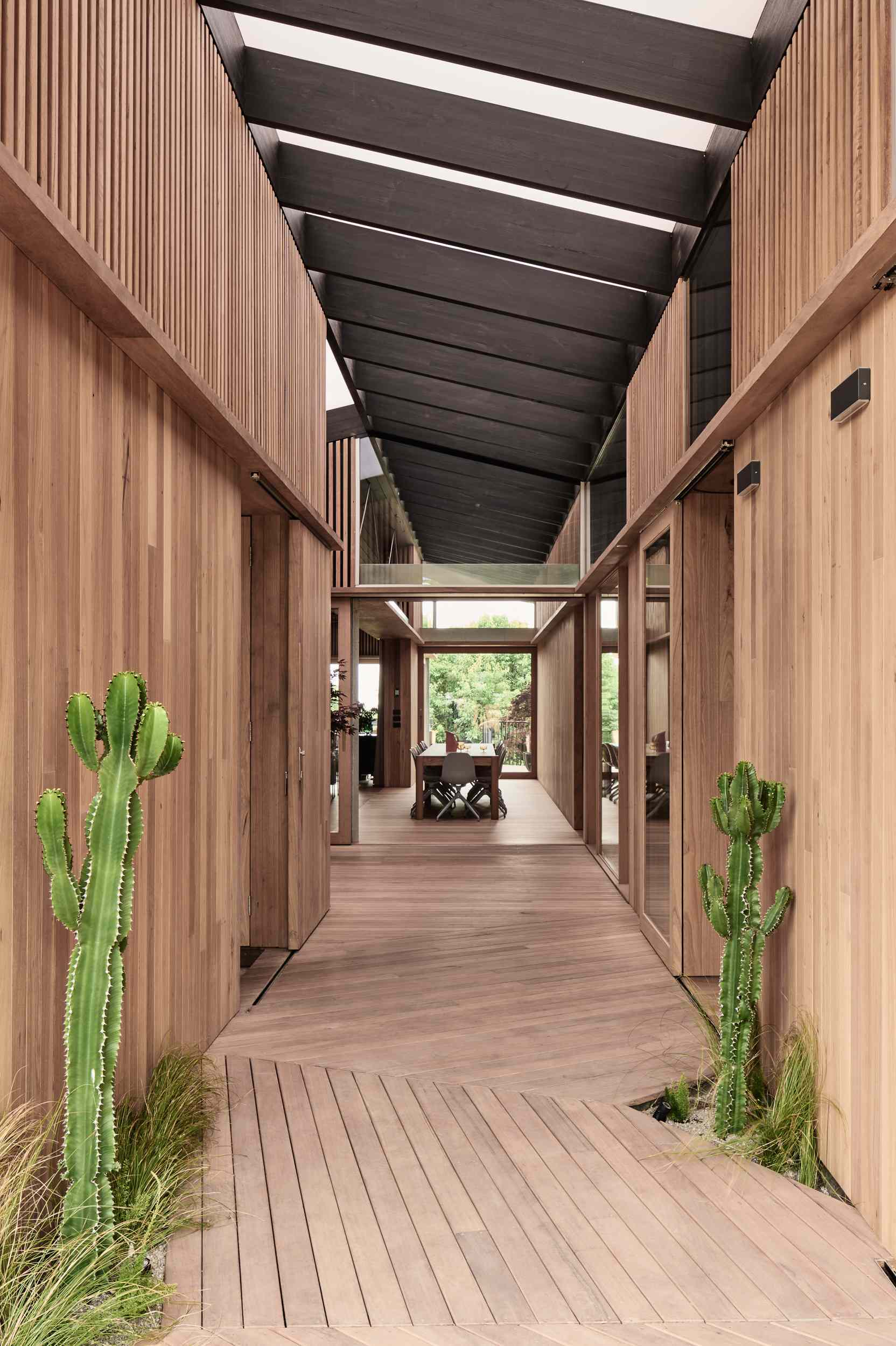 The interior boardwalk flows straight into the dining area, where timber walls and flooring create a warm, cohesive feel. A long wooden table anchors the space, paired with soft grey chairs that add a gentle contrast to the rich natural tones.
