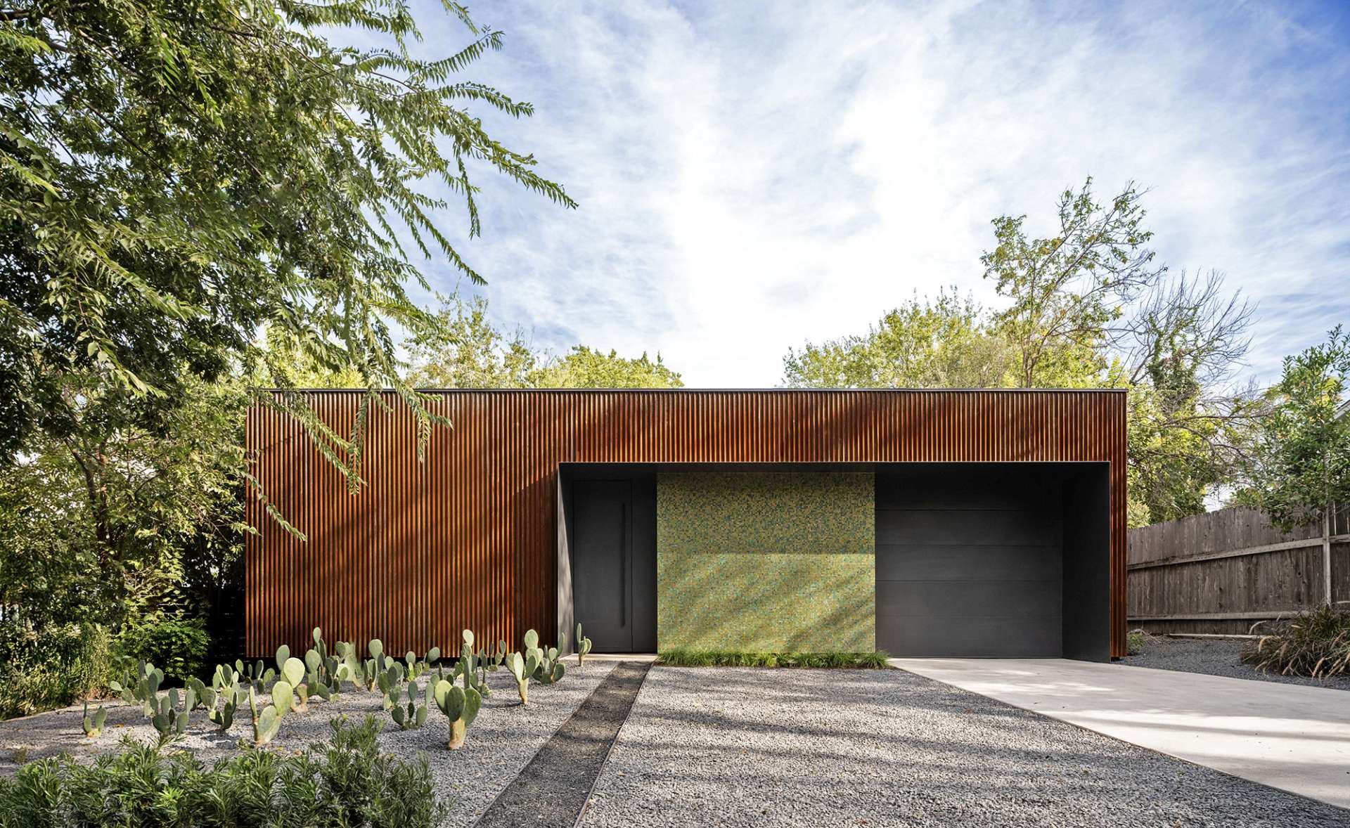A bold mix of industrial and refined materials, this modern home pairs rusted corrugated steel with a shimmering mosaic wall for a striking first impression. The narrow window offers a glimpse into the tranquil, glass-lined interior.