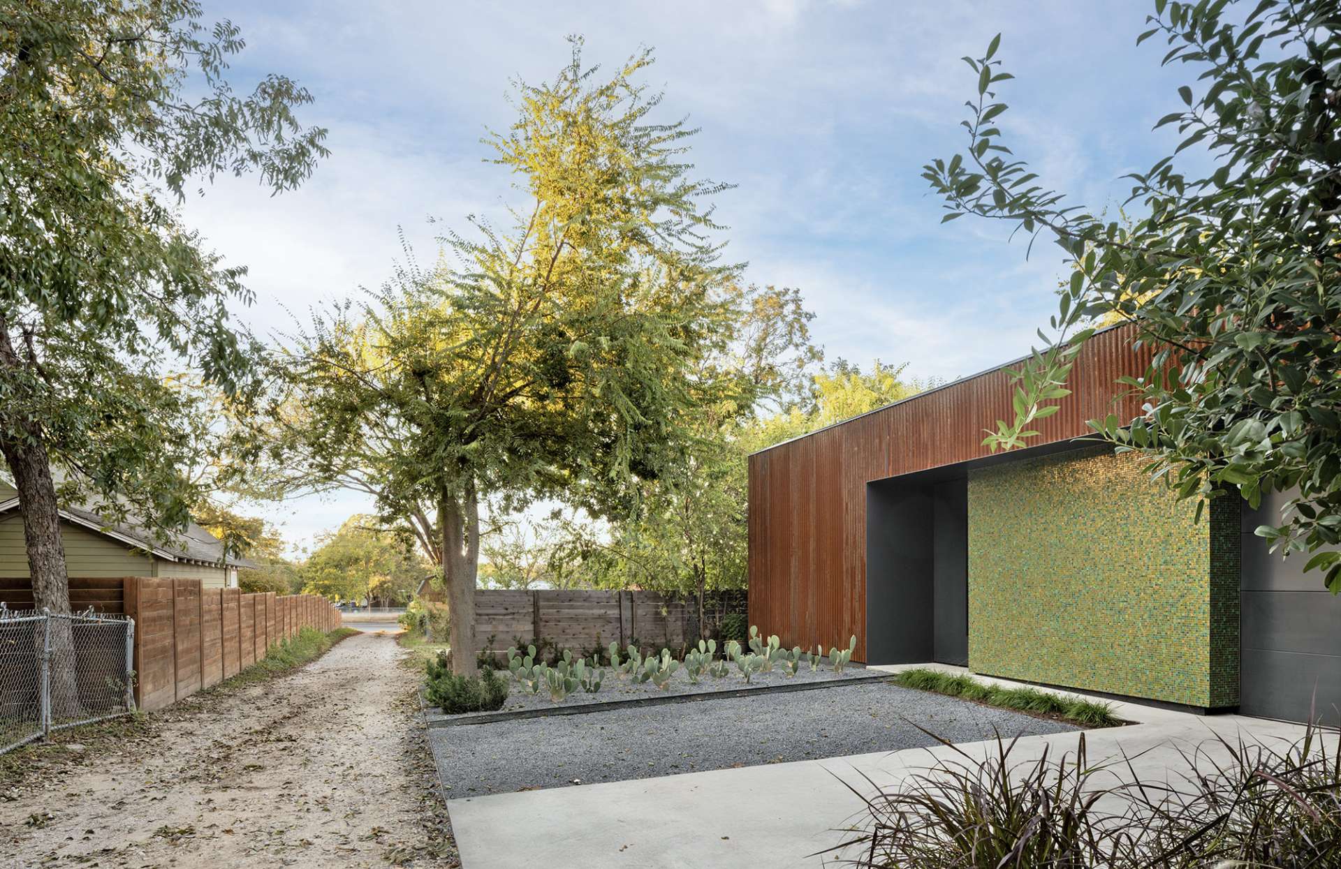 A bold mix of industrial and refined materials, this modern home pairs rusted corrugated steel with a shimmering mosaic wall for a striking first impression. The narrow window offers a glimpse into the tranquil, glass-lined interior.