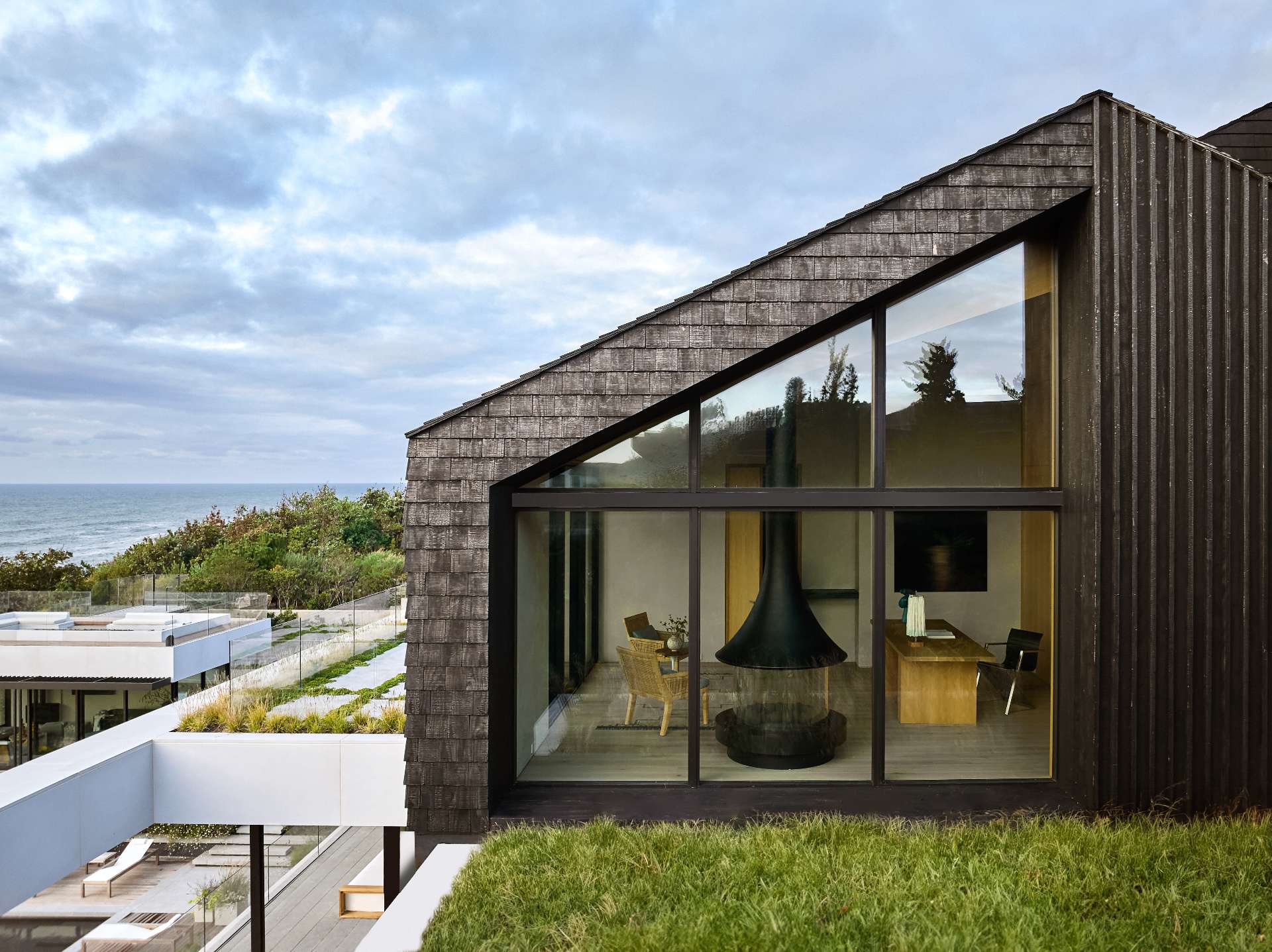 Framed by a sharply pitched roof and clad in wood shingles, the expansive floor-to-ceiling windows serve as a striking visual anchor, seamlessly connecting the interior living space to the surrounding landscape. The angled glass panels echo the geometry of the architecture, while offering unobstructed views of the ocean and sky beyond.