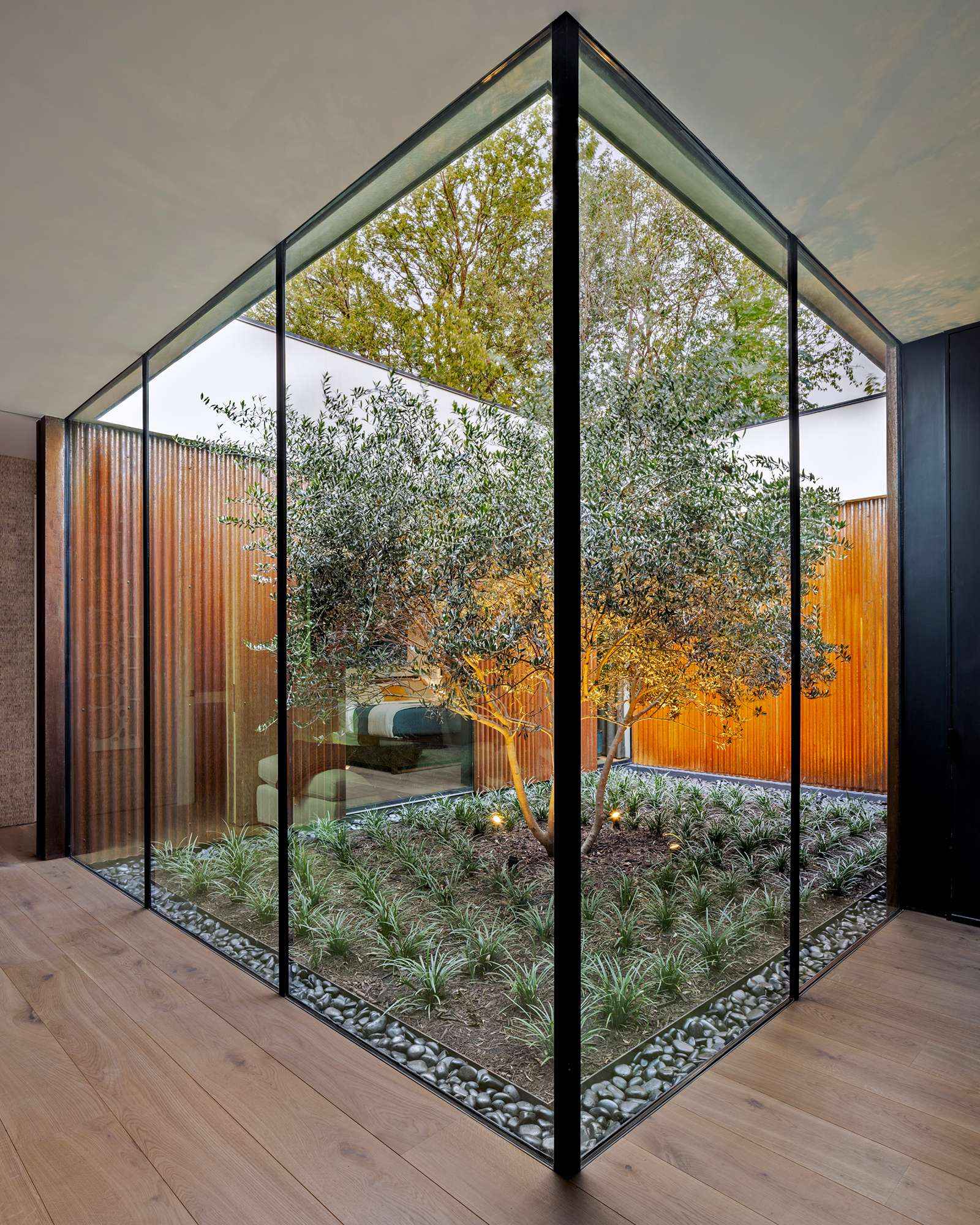 This serene courtyard anchors the home with a sculptural olive tree framed by floor-to-ceiling glass. Natural light floods the kitchen and living spaces while rusted steel cladding brings warmth and texture to the modern design.