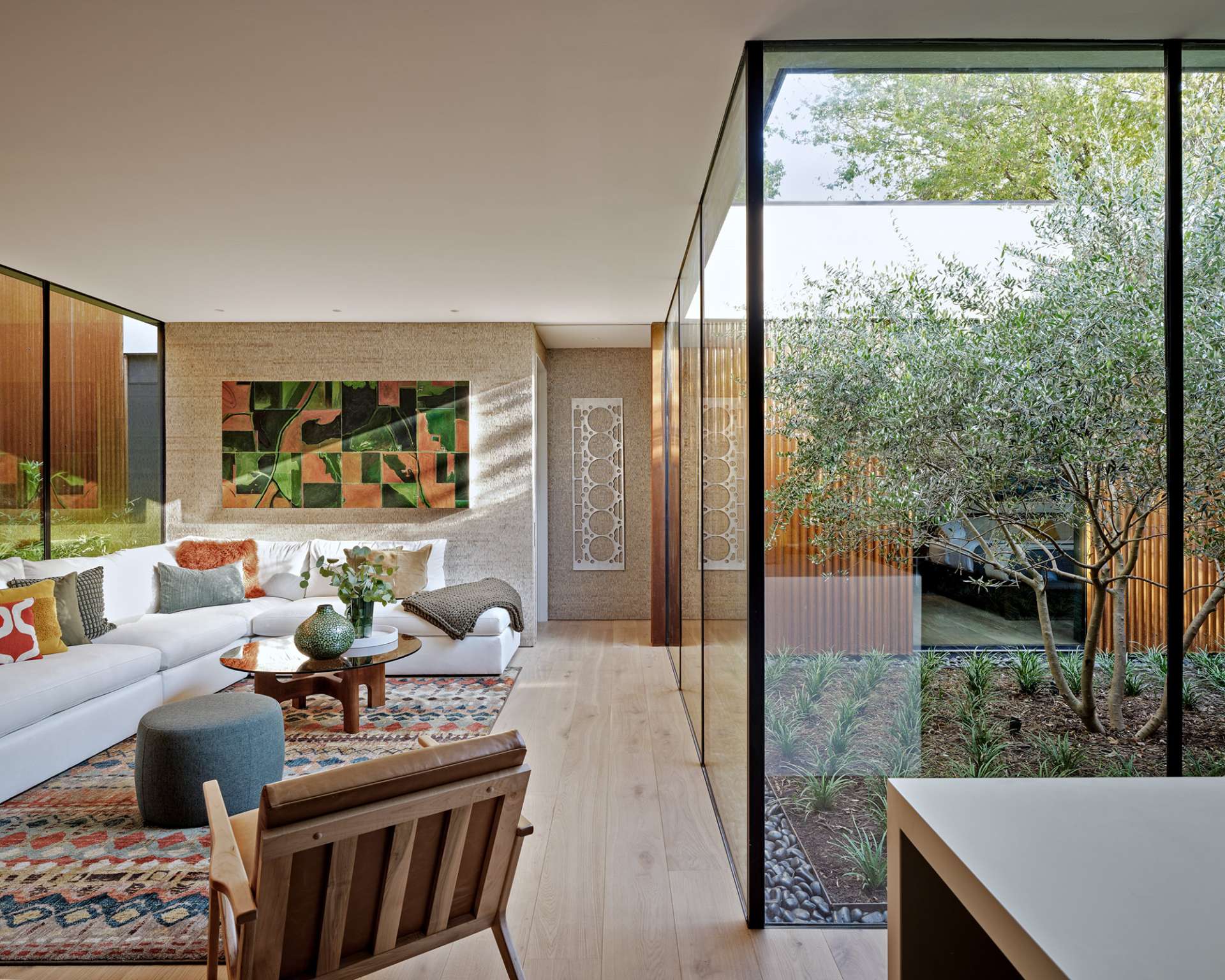 This cozy living room blends earthy textures and warm wood tones with expansive glass walls that open to lush bamboo landscaping. A colorful rug and sculptural furnishings add comfort and character to the modern minimalist space.