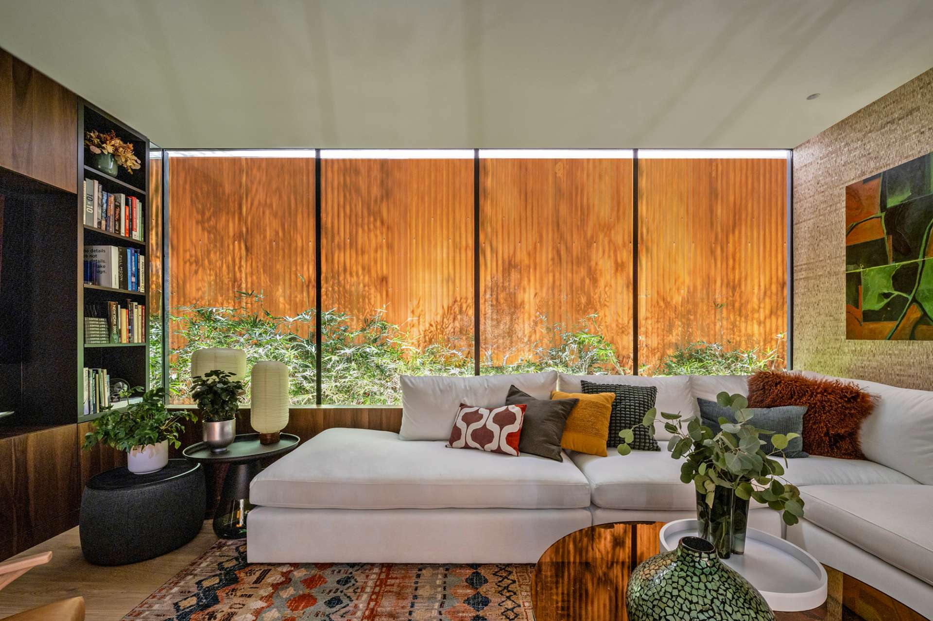 This cozy living room blends earthy textures and warm wood tones with expansive glass walls that open to lush bamboo landscaping. A colorful rug and sculptural furnishings add comfort and character to the modern minimalist space.