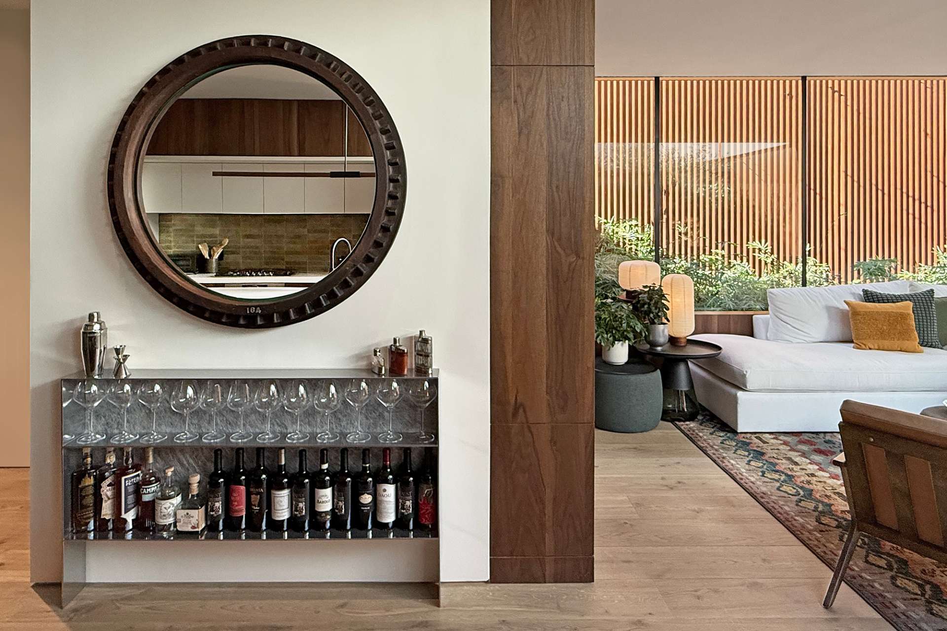 A metal bar unit displays wine bottles and glassware with effortless style, paired with a sculptural round mirror. Warm wood finishes and soft textures in the background create a cozy yet refined living space.