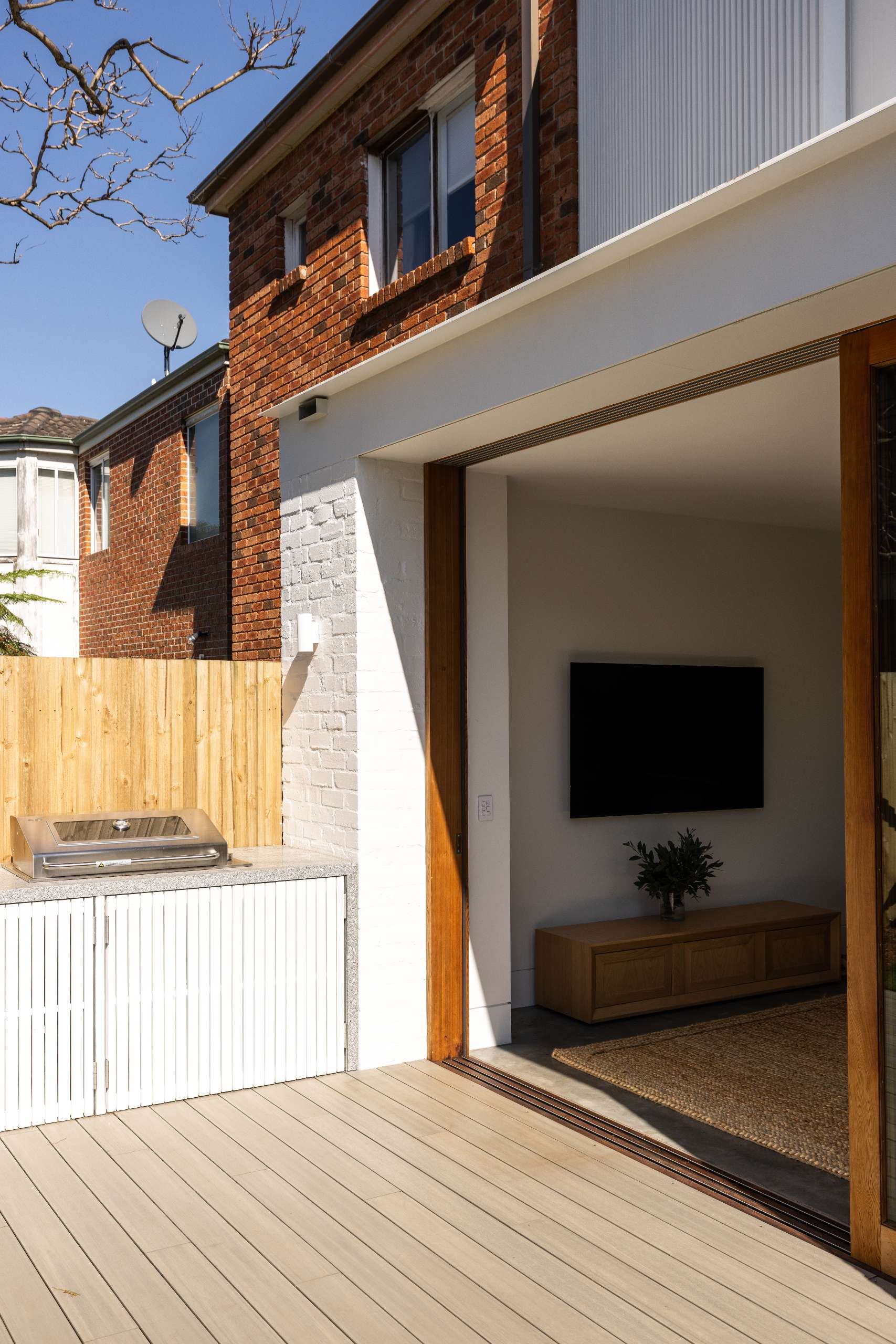 A sleek outdoor BBQ kitchen sits against a timber fence, featuring clean white battens with hidden storage, seamlessly extending from an open-plan living area onto a light timber deck.