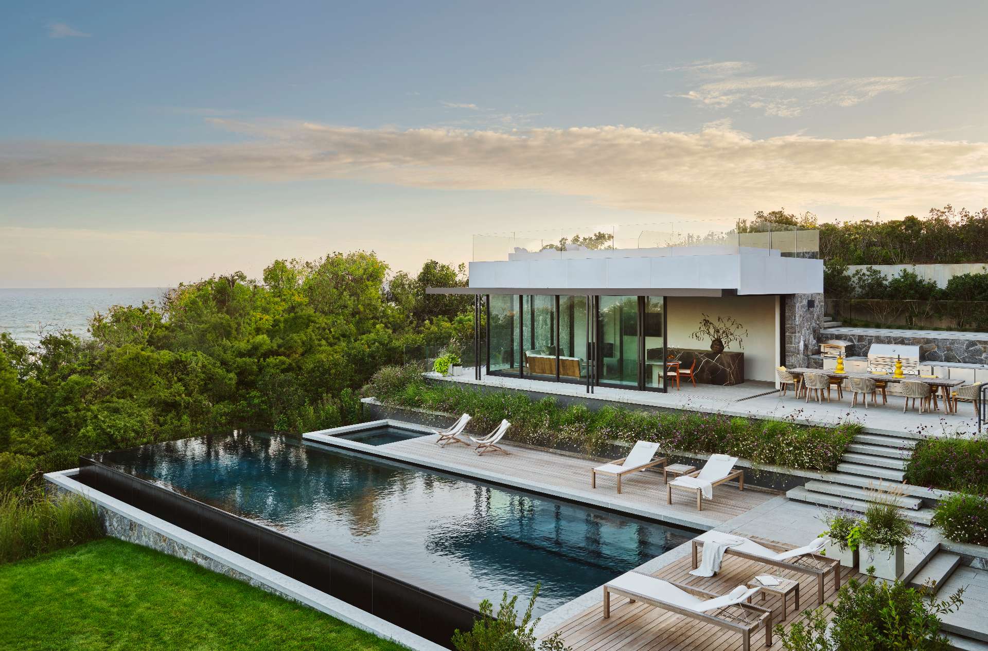 With panoramic ocean views as its backdrop, the lower terrace of this modern house is anchored by an infinity-edge pool and flanked by generous lounge areas. A glass-walled pool house opens onto the terrace, while a built-in BBQ area nearby invites outdoor entertaining.