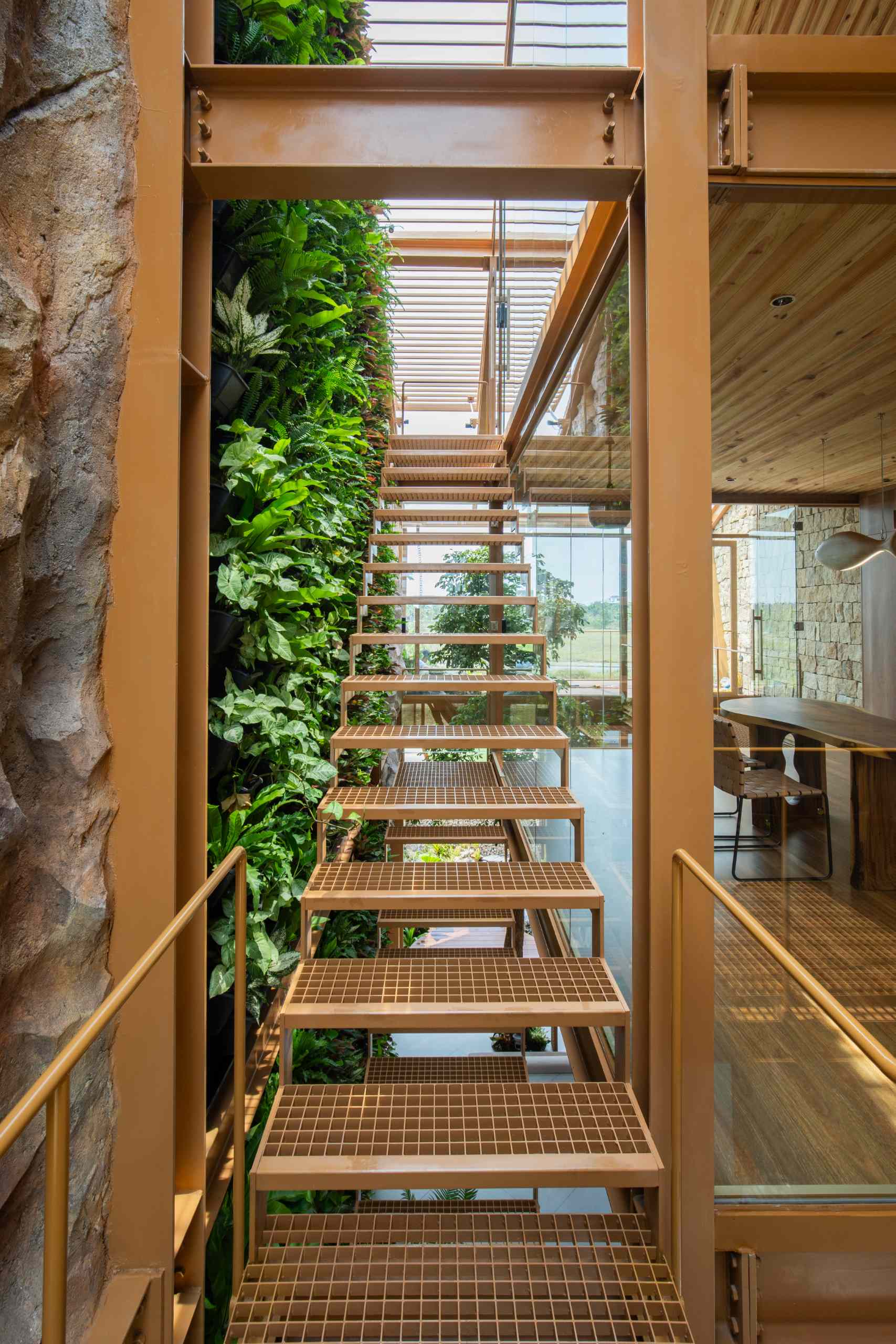 Steel stairs with an open-grid design bring an industrial edge while keeping things light and airy. One side frames a lush vertical garden, the other opens to natural stone and glass, blending raw materials with greenery and light.