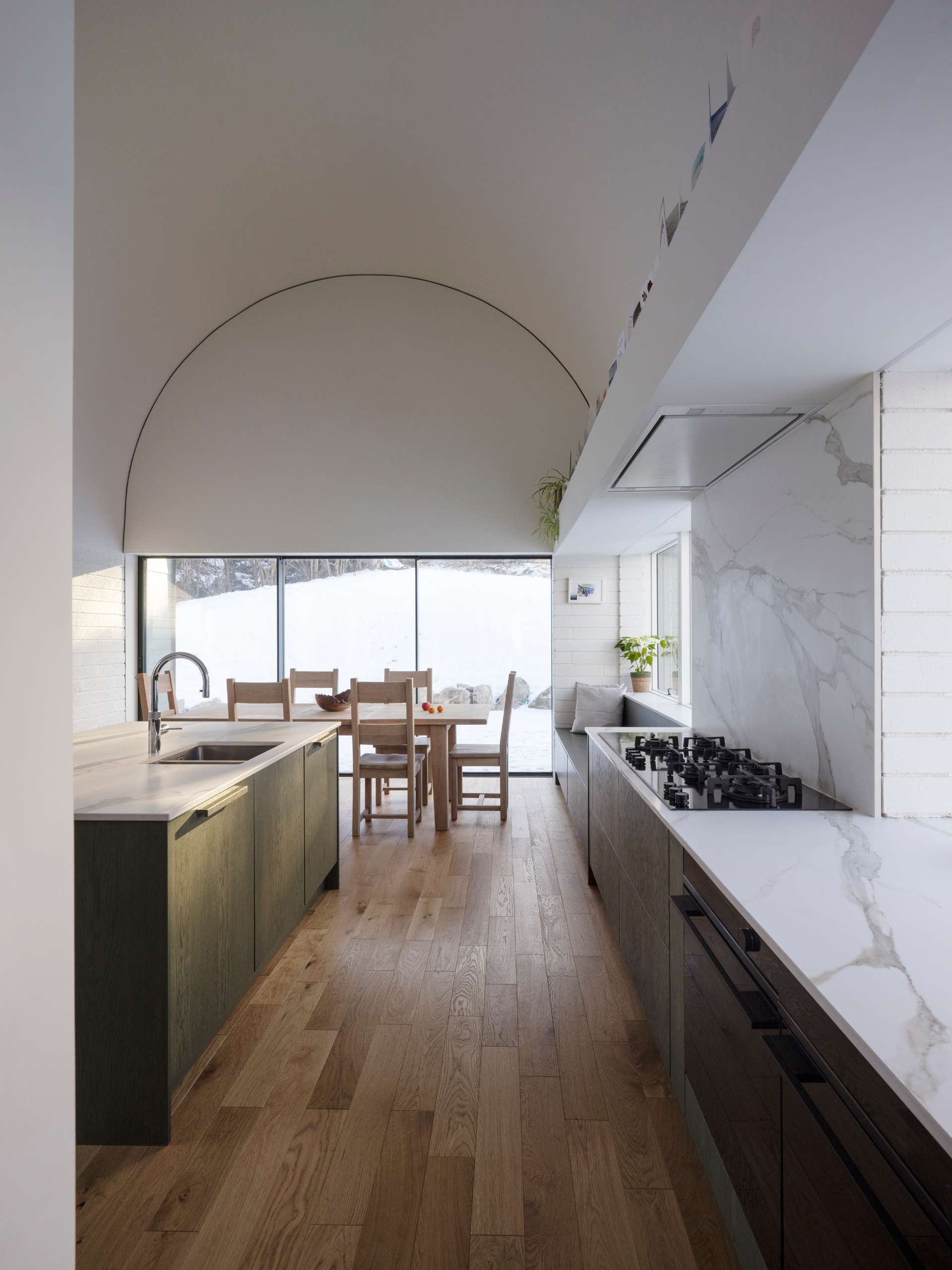 This serene kitchen pairs a vaulted ceiling with natural oak floors and marble countertops for a quietly luxurious feel. Floor-to-ceiling glass opens the space to snowy views, while warm wood tones and built-in seating create a calm, considered atmosphere that’s both functional and beautiful.