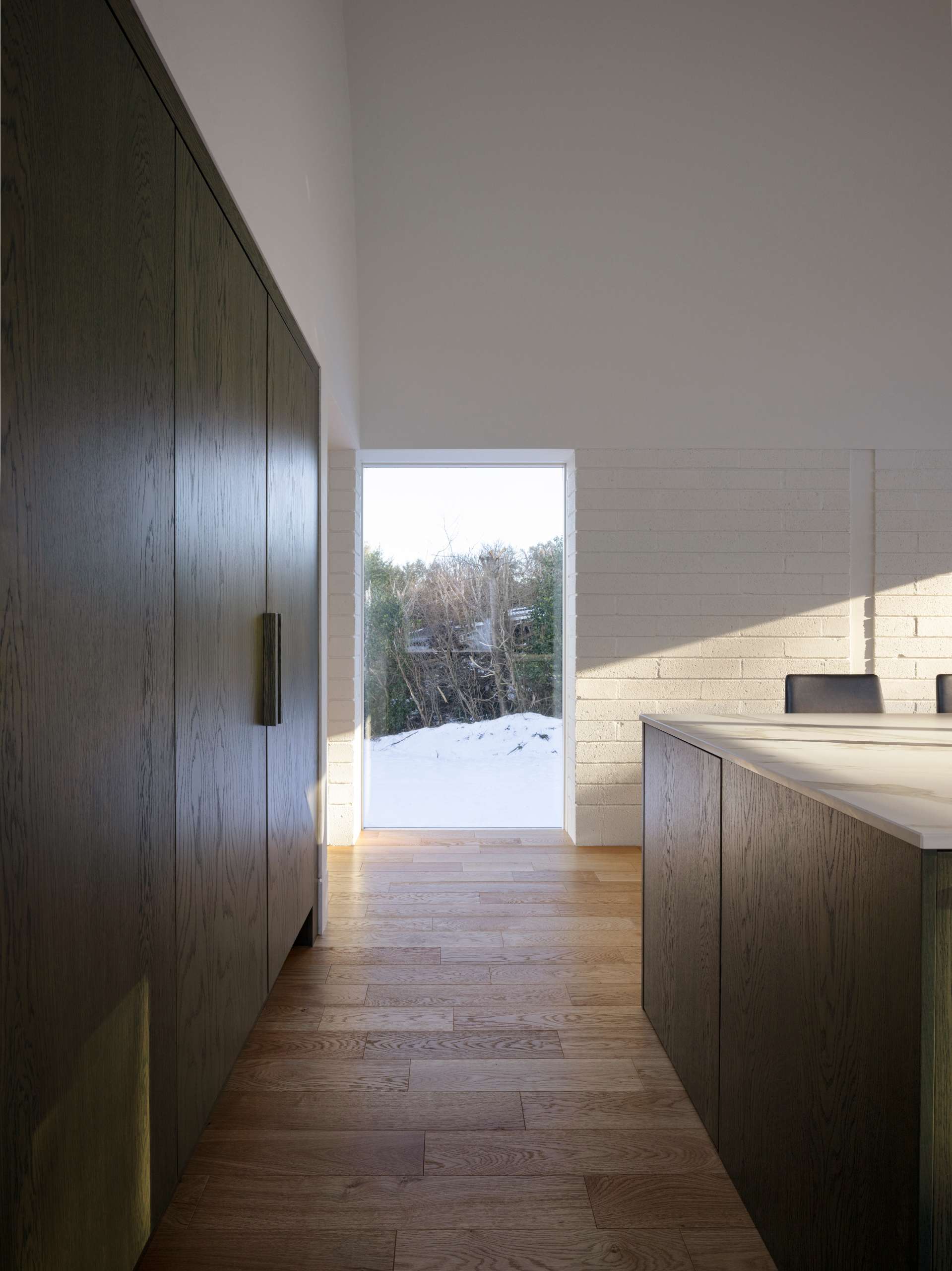 This serene kitchen pairs a vaulted ceiling with natural oak floors and marble countertops for a quietly luxurious feel. Floor-to-ceiling glass opens the space to snowy views, while warm wood tones and built-in seating create a calm, considered atmosphere that’s both functional and beautiful.