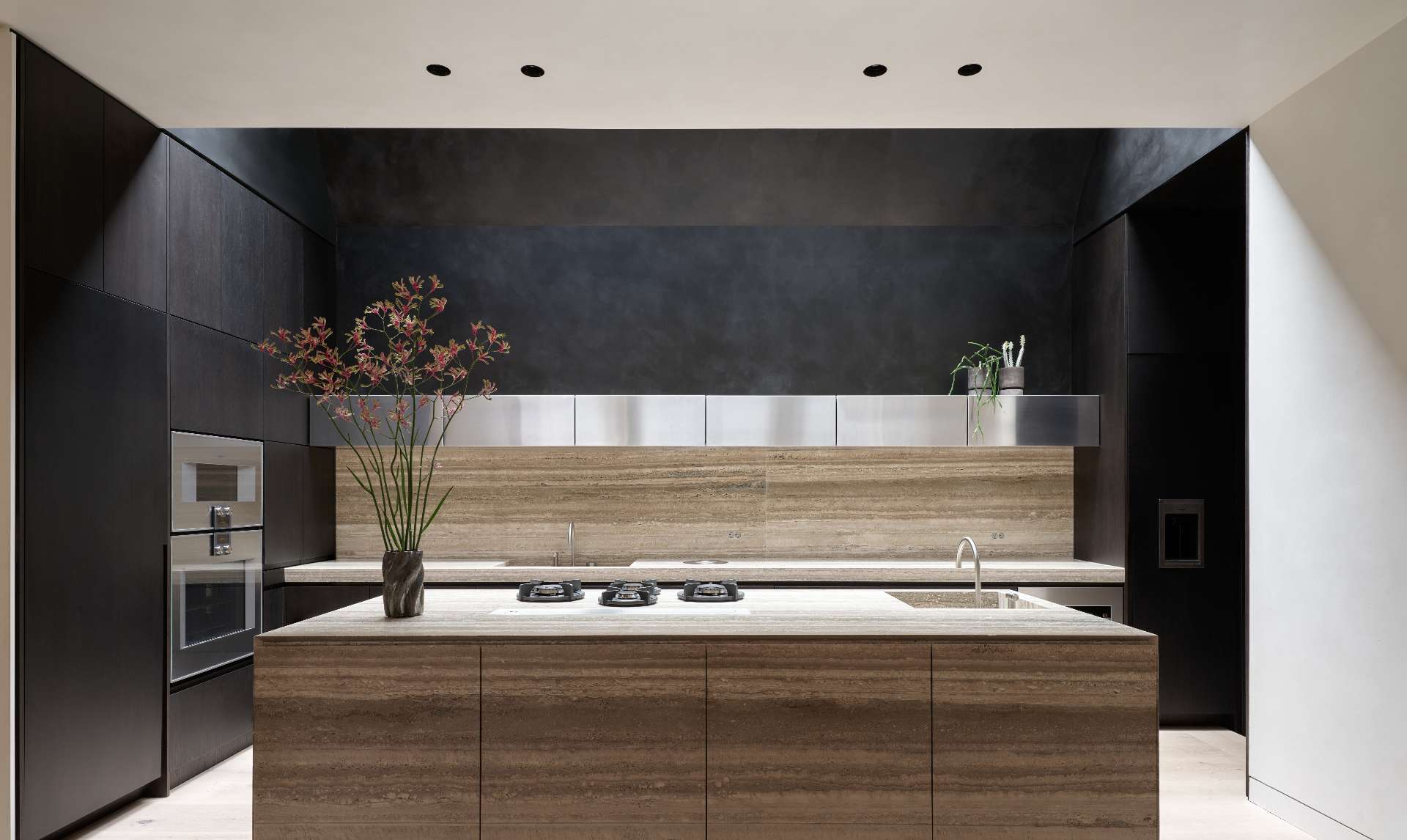 The kitchen pairs dark walnut cabinetry with wide-plank white oak floors. A travertine island and matching backsplash anchors the space, while a skylight above draws in natural light, emphasizing the material contrasts and continuing the home’s shift from intimate to expansive volumes.