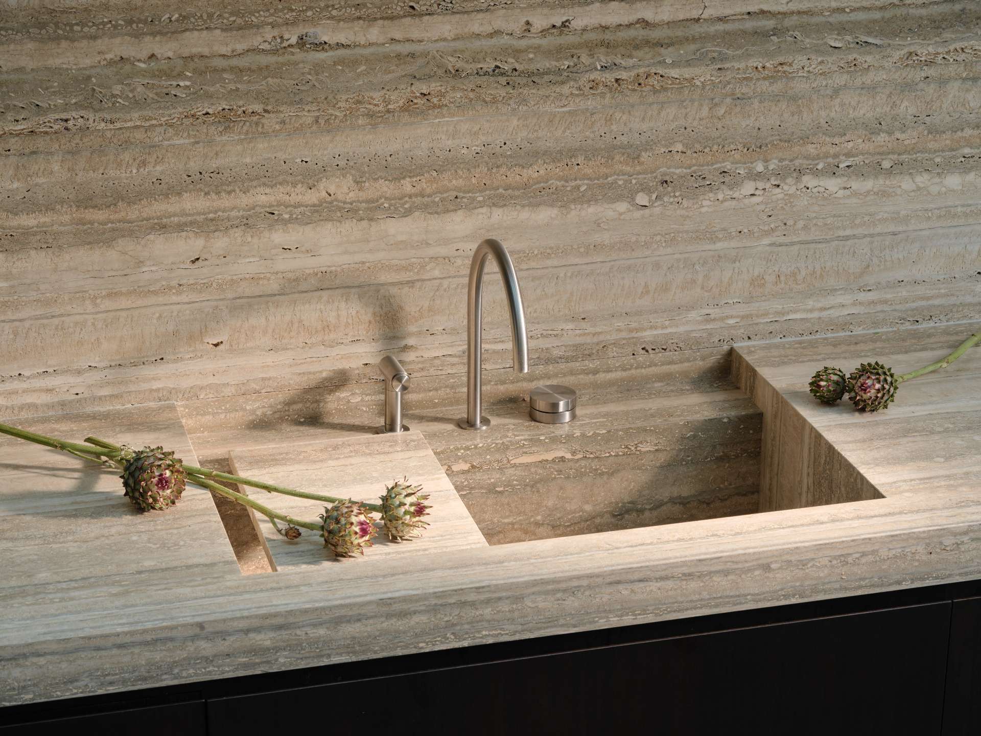 The kitchen pairs dark walnut cabinetry with wide-plank white oak floors. A travertine island and matching backsplash anchors the space, while a skylight above draws in natural light, emphasizing the material contrasts and continuing the home’s shift from intimate to expansive volumes.