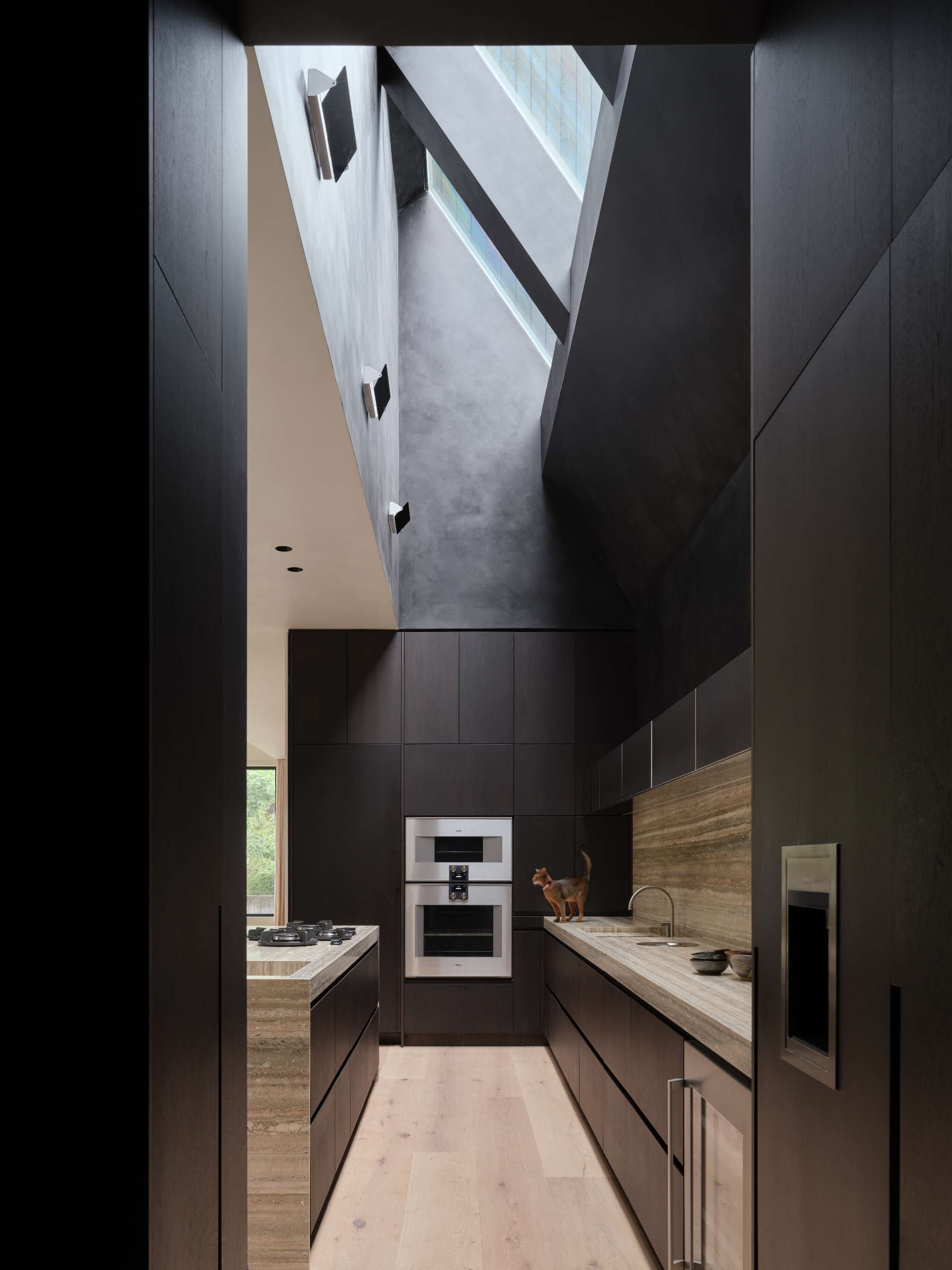 The kitchen pairs dark walnut cabinetry with wide-plank white oak floors. A travertine island and matching backsplash anchors the space, while a skylight above draws in natural light, emphasizing the material contrasts and continuing the home’s shift from intimate to expansive volumes.