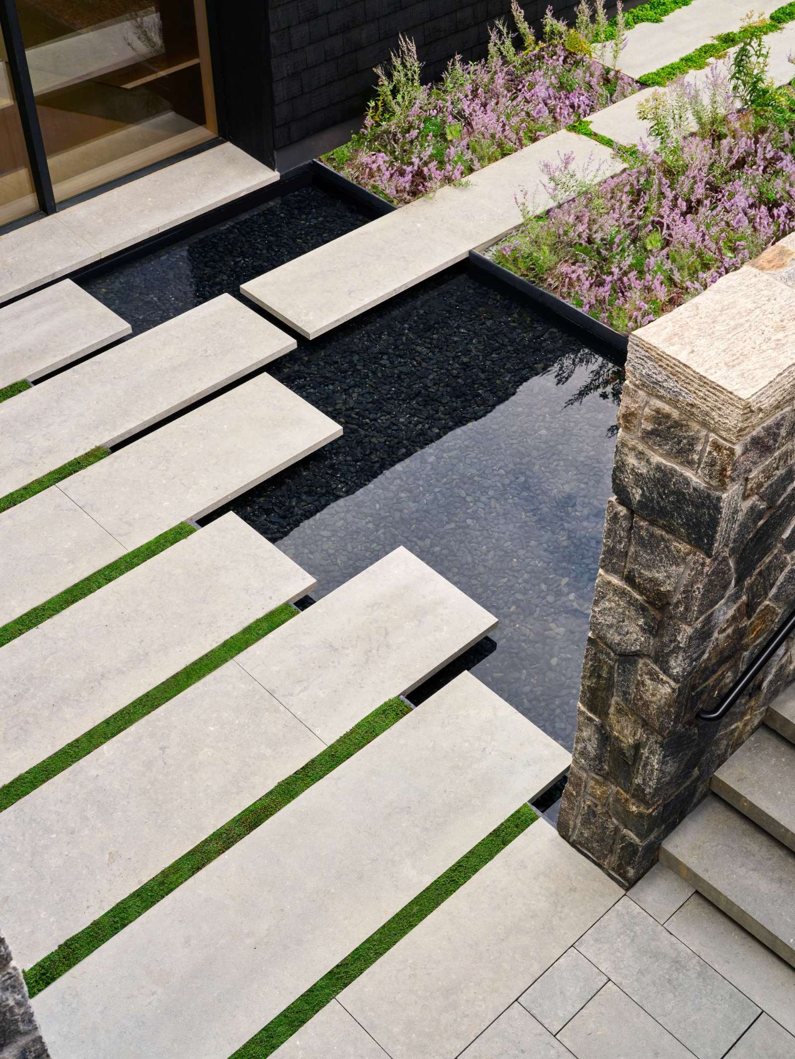 Stepping down towards the home, large pavers appear to float above a shallow reflective pool, framed by soft green moss that adds rhythm and contrast. Rugged stone walls anchor the space with texture and warmth, while the vertical wood slats filters sunlight and casts dynamic shadows.