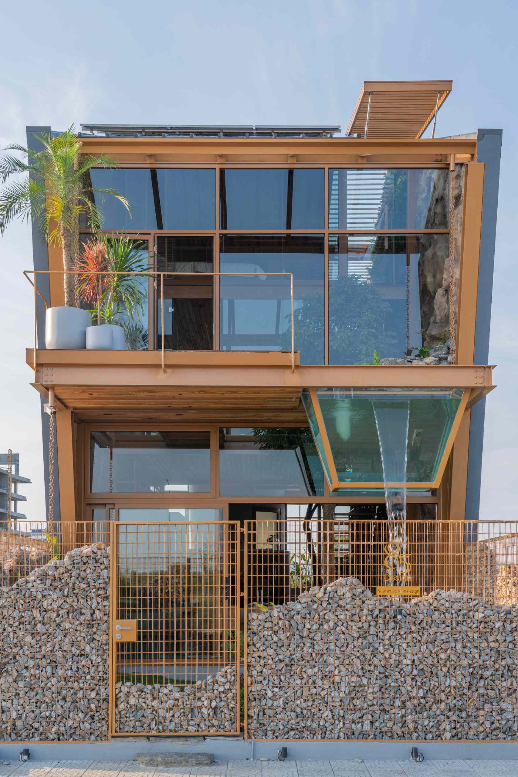 The exterior of this live-work building blends modern lines with natural materials, steel framing, warm wood, and wide glass panels. A glass box extending from the upper floor doubles as a water feature, while a stone-filled fence below adds texture and an earthy contrast.
