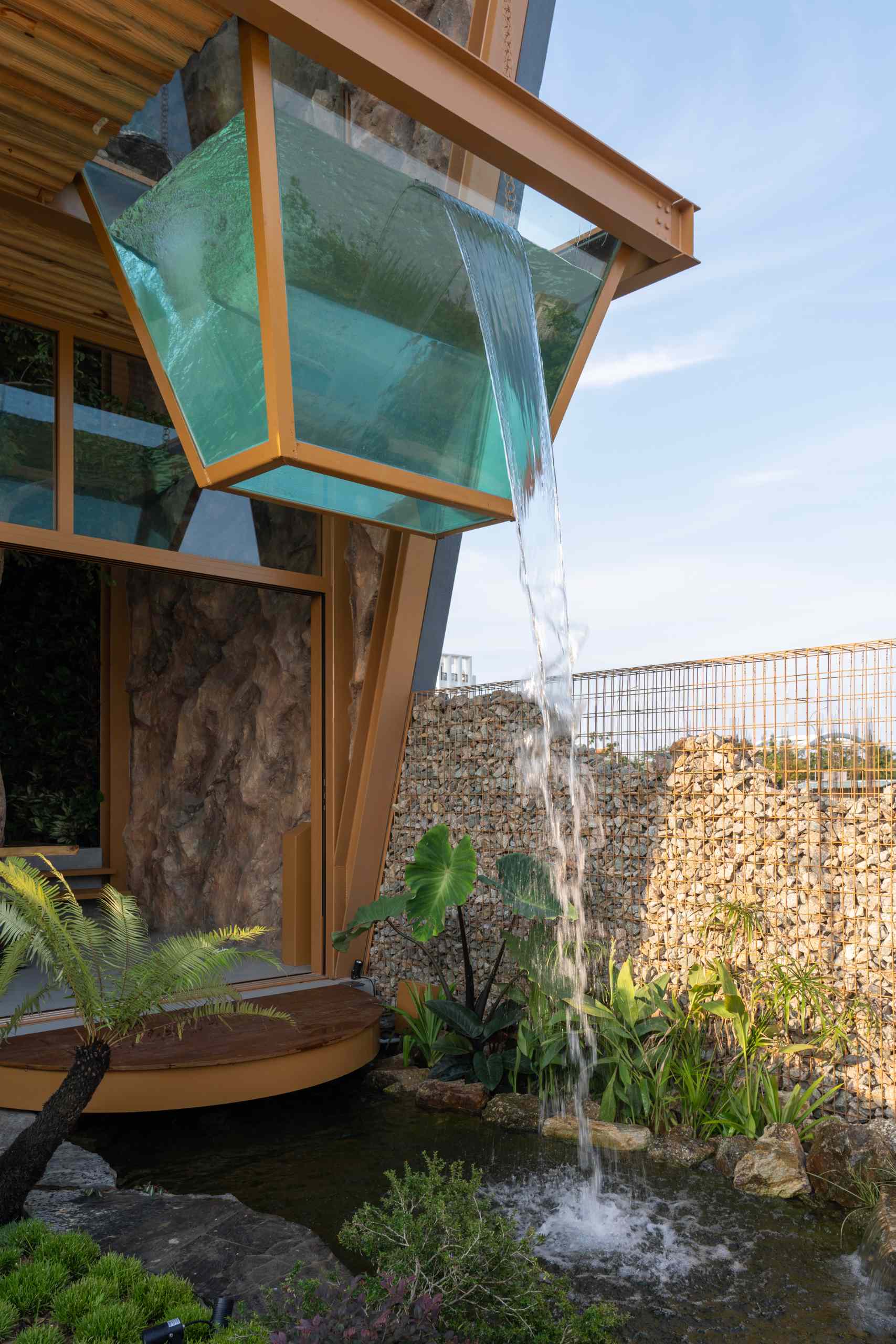 A glass volume suspended above a pond creates a striking waterfall that cools the space and adds a calming sound. Surrounded by tropical plants, natural stone, and a textured gabion wall, the garden feels like a small, living ecosystem built into the heart of a modern home.