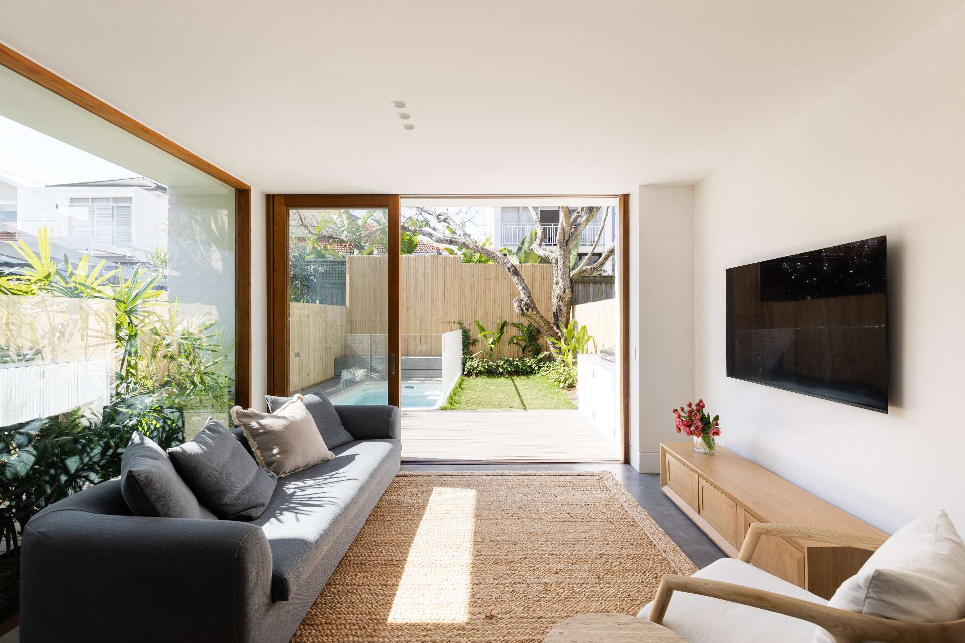 This living space is open-plan and visually expansive, with floor-to-ceiling glass doors, a low grey sofa, and a woven jute rug. Timber accents and white walls create a calm, minimalist atmosphere.