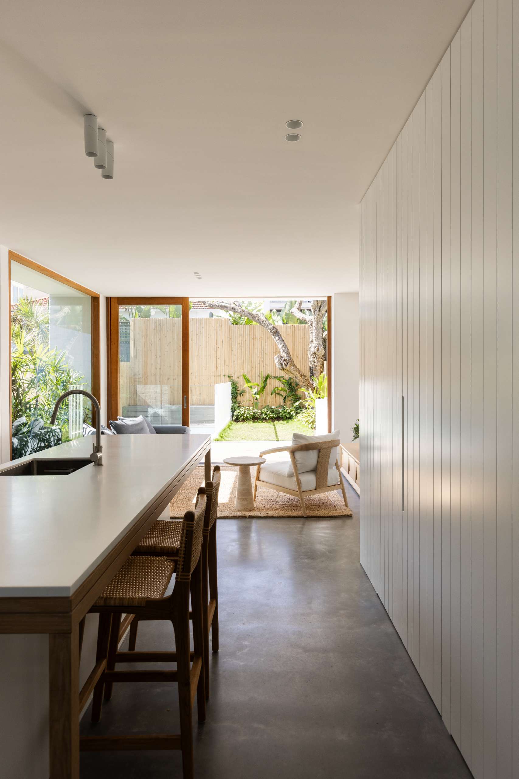 This living space is open-plan and visually expansive, with floor-to-ceiling glass doors, a low grey sofa, and a woven jute rug. Timber accents and white walls create a calm, minimalist atmosphere.