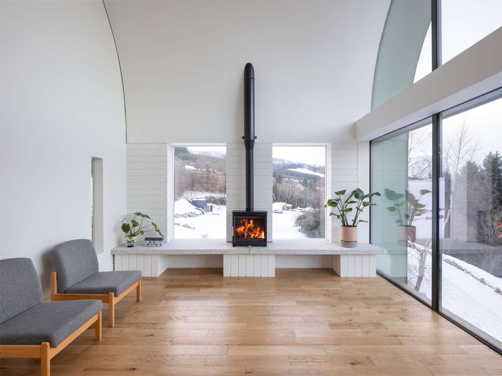A wood-burning stove sits at the center of one living room wall, framed by crisp white brick and warm timber floors. Simple furnishings and natural textures create a calm, inviting atmosphere that feels both modern and rooted in place.
