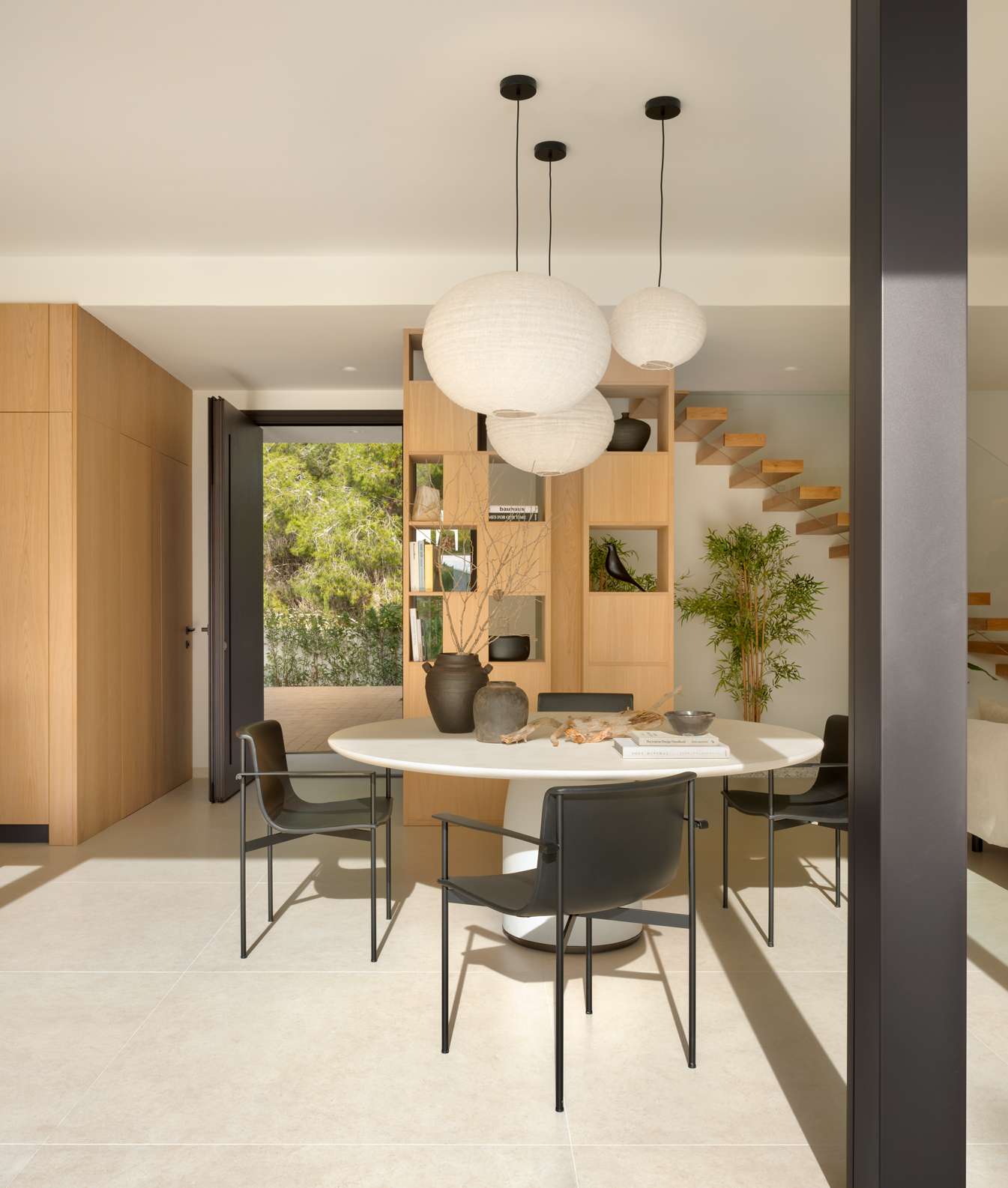 A soft sculptural palette defines this serene dining space, where paper lanterns hover gently above a round pedestal table. Built-in timber shelving adds visual warmth and a place for personal touches, while black chairs and ceramic accents introduce a sense of balance and contrast.