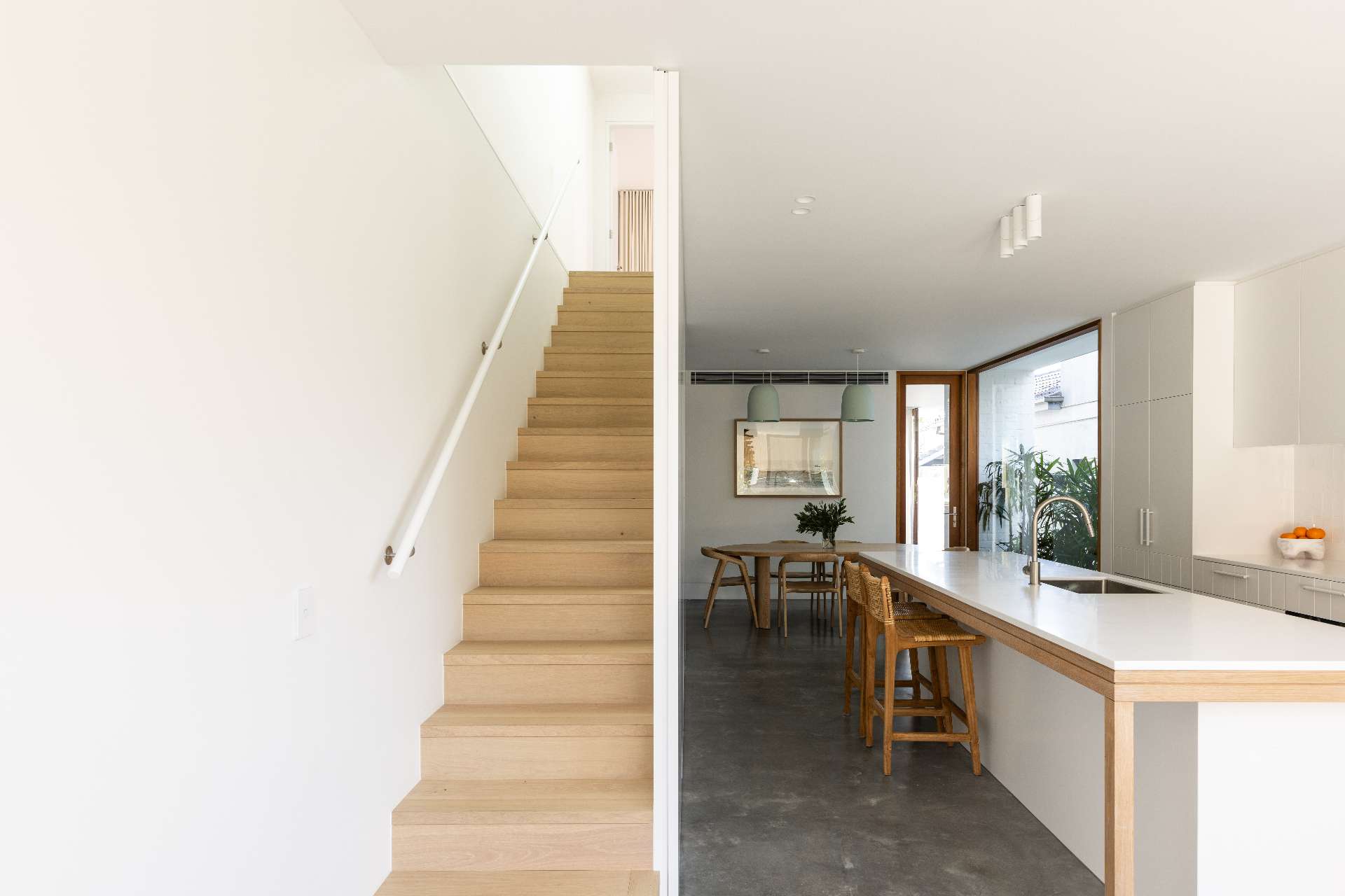 A soft minimalist kitchen defined by matte white cabinetry, integrated appliances, and a long timber-accented island. Woven stools add warmth and texture, while hidden storage beneath the staircase keeps the space feeling clean and refined.