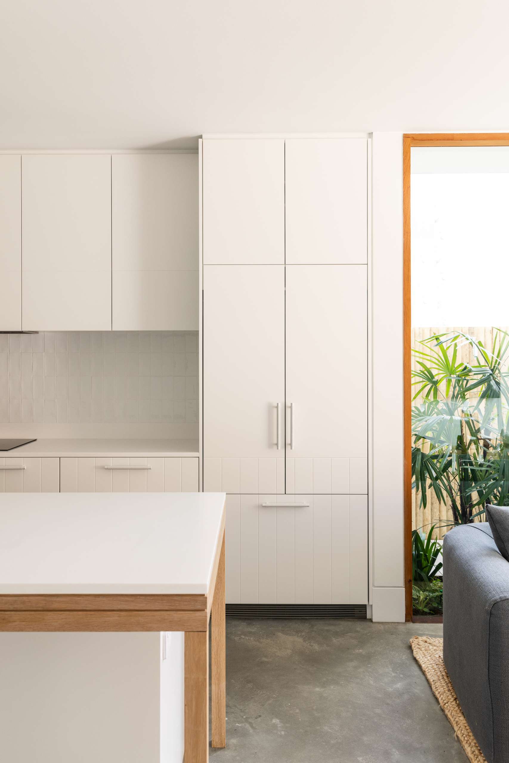 A soft minimalist kitchen defined by matte white cabinetry, integrated appliances, and a long timber-accented island. Woven stools add warmth and texture, while hidden storage beneath the staircase keeps the space feeling clean and refined.