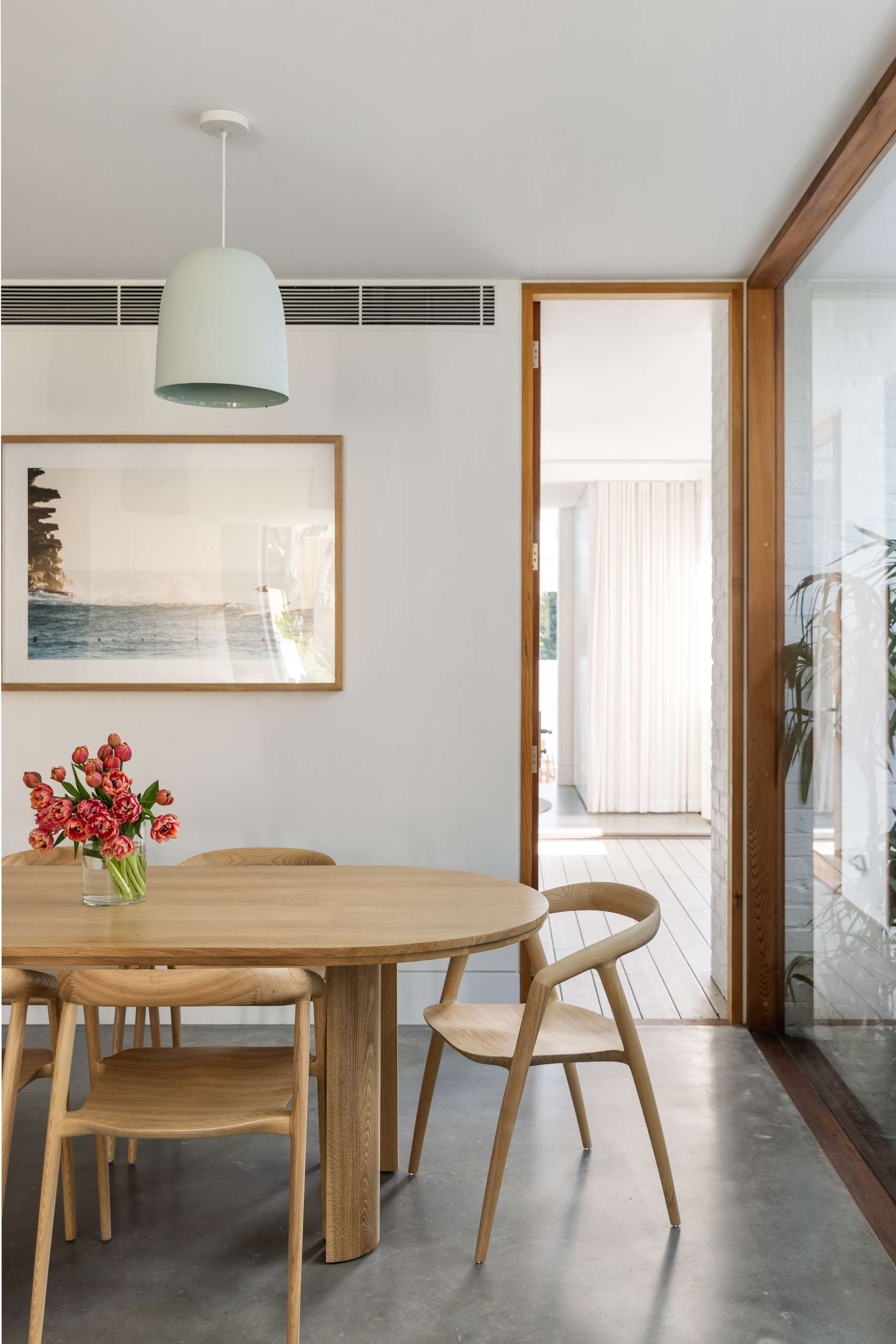 A serene dining space that flows from the kitchen, defined by coastal minimalism. Sculptural timber chairs surround a rounded table, while soft mist-hued pendants and a framed ocean print bring in subtle beachside charm.
