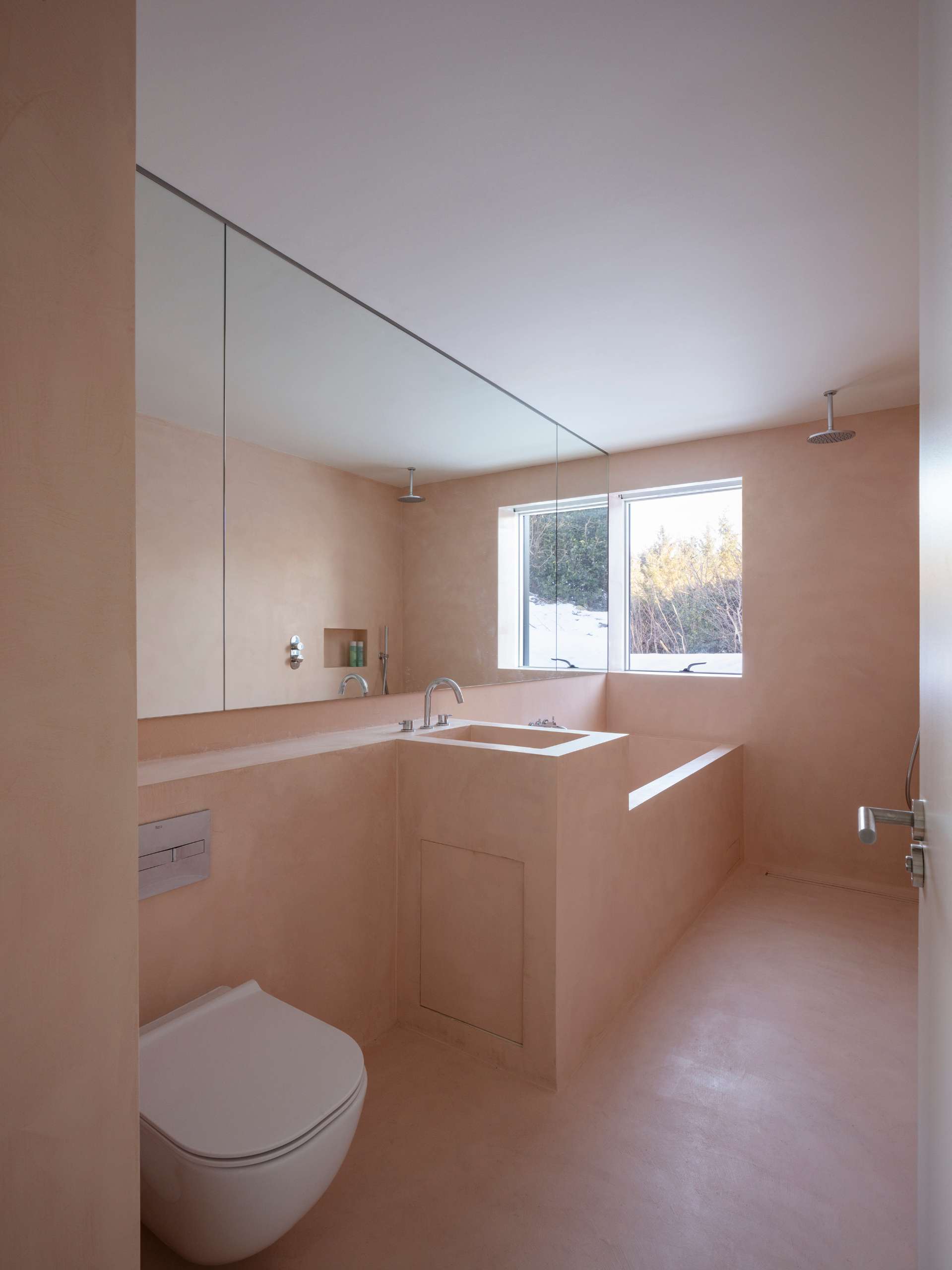 In this modern bathroom, there's a peach-toned finish that creates a warm, monochromatic aesthetic from floor to ceiling. Built-in elements, including the vanity and tub, enhance the clean lines and architectural simplicity. A full-width mirror and window fill the space with light, making it feel open and calming.
