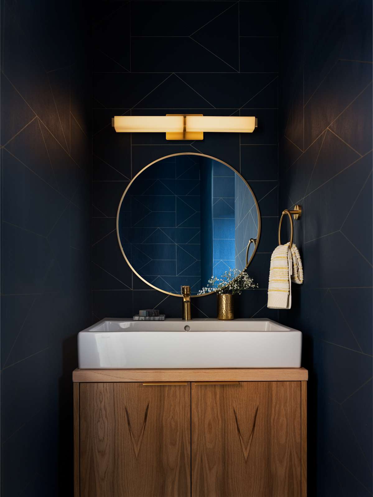 A sophisticated powder room with a moody navy backdrop has subtle geometric lines for added depth. A round brass mirror and matching fixtures bring a touch of glam, while the warm wood vanity adds natural texture. The modern light fixture and minimalist styling create a refined, spa-like vibe in a small space.