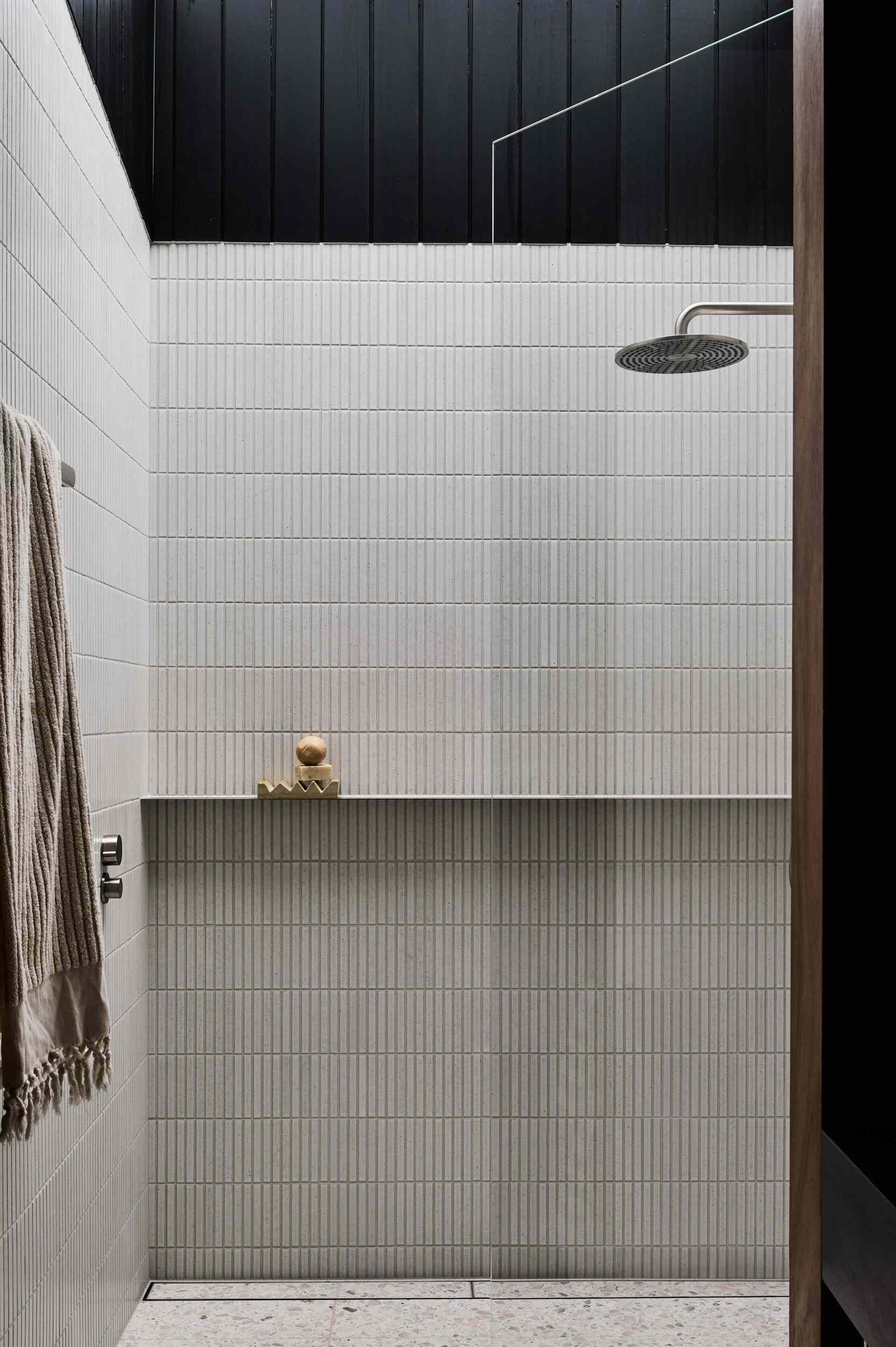 This shower features slim, vertically ridged off-white tiles that create a sense of height and texture. A sleek built-in ledge provides minimalist storage, while the rain showerhead adds a modern, spa-like touch.