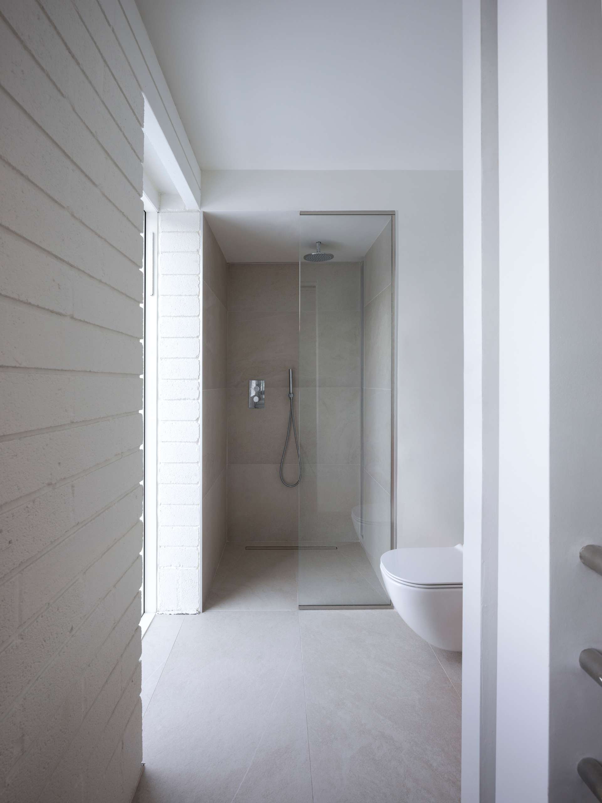 In this modern bathroom, there's soft neutral tones and a calming, uncluttered feel. A walk-in shower with a glass enclosure keeps the space light and open, while the floating toilet adds to the streamlined look. Subtle texture from white painted brick contrasts beautifully with the smooth tile surfaces.