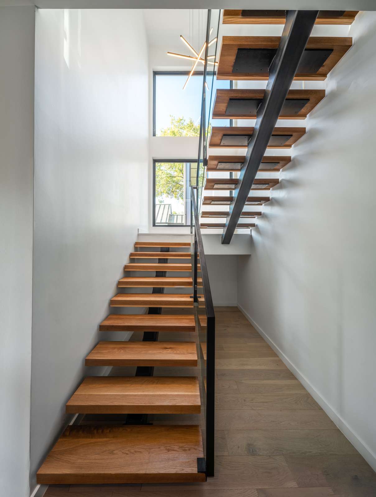 Striking modern staircase design featuring floating wood treads with black steel supports for a clean, architectural look. Large windows fill the stairwell with natural light, creating a bright and open feel. The minimalist handrail and sculptural pendant light add a sleek finishing touch. Perfect inspiration for anyone remodeling a contemporary home with an eye for bold design and natural materials.