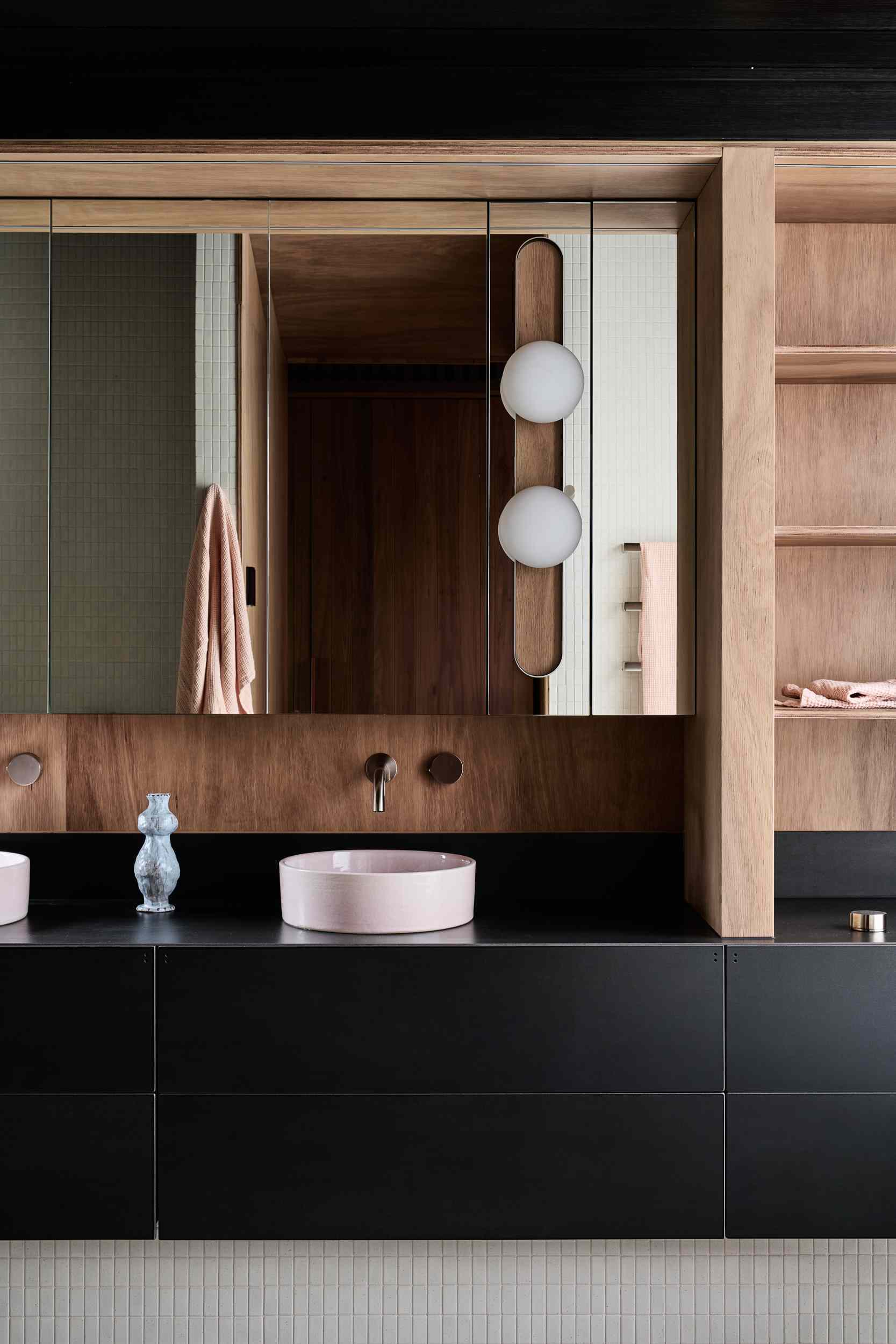 This bathroom pairs natural timber with matte black cabinetry for a warm, modern contrast. Soft pink basins and sculptural lighting add a refined, playful touch. A freestanding tub sits by sheer curtains, inviting calm and natural light into the minimalist space.