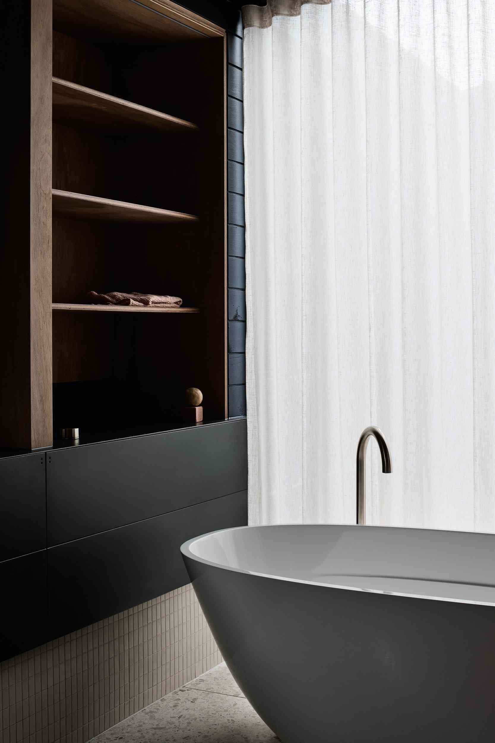 This bathroom pairs natural timber with matte black cabinetry for a warm, modern contrast. Soft pink basins and sculptural lighting add a refined, playful touch. A freestanding tub sits by sheer curtains, inviting calm and natural light into the minimalist space.