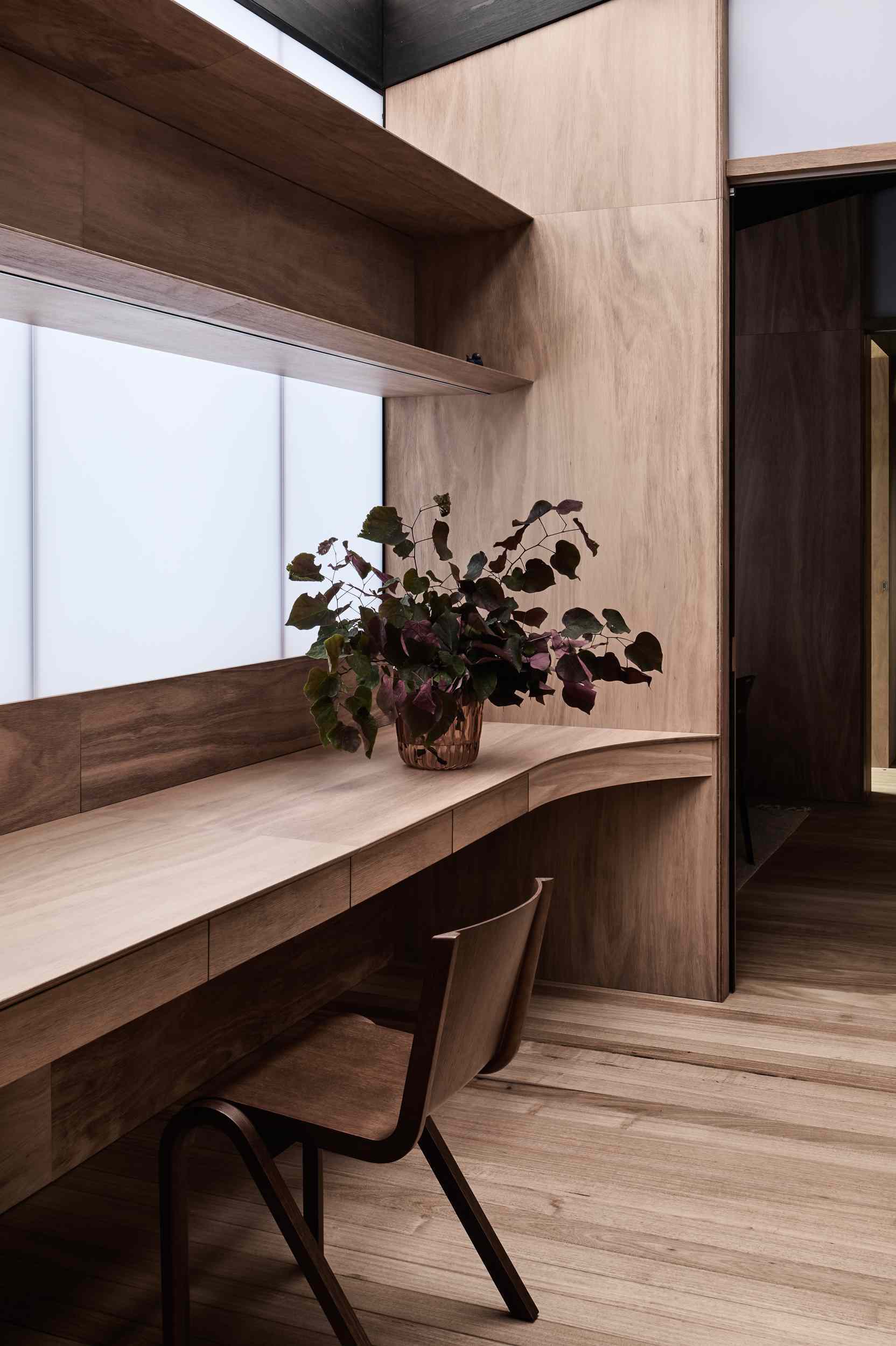 A custom built-in desk flows seamlessly into warm timber walls and cabinetry. Frosted glazing softly diffuses light, while a wood chair and potted plant add a quiet elegance to the minimalist space.