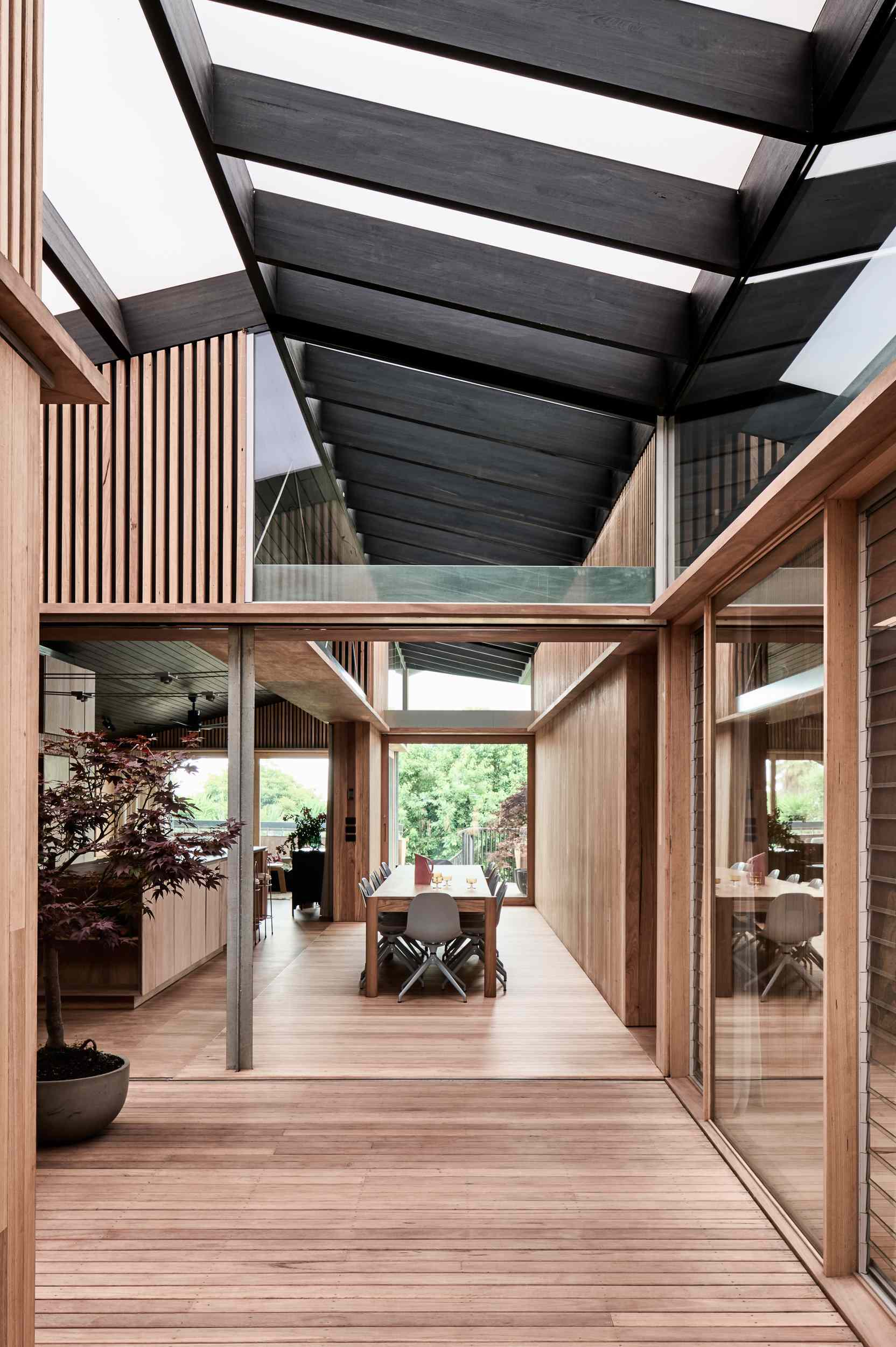 The interior boardwalk flows straight into the dining area, where timber walls and flooring create a warm, cohesive feel. A long wooden table anchors the space, paired with soft grey chairs that add a gentle contrast to the rich natural tones.