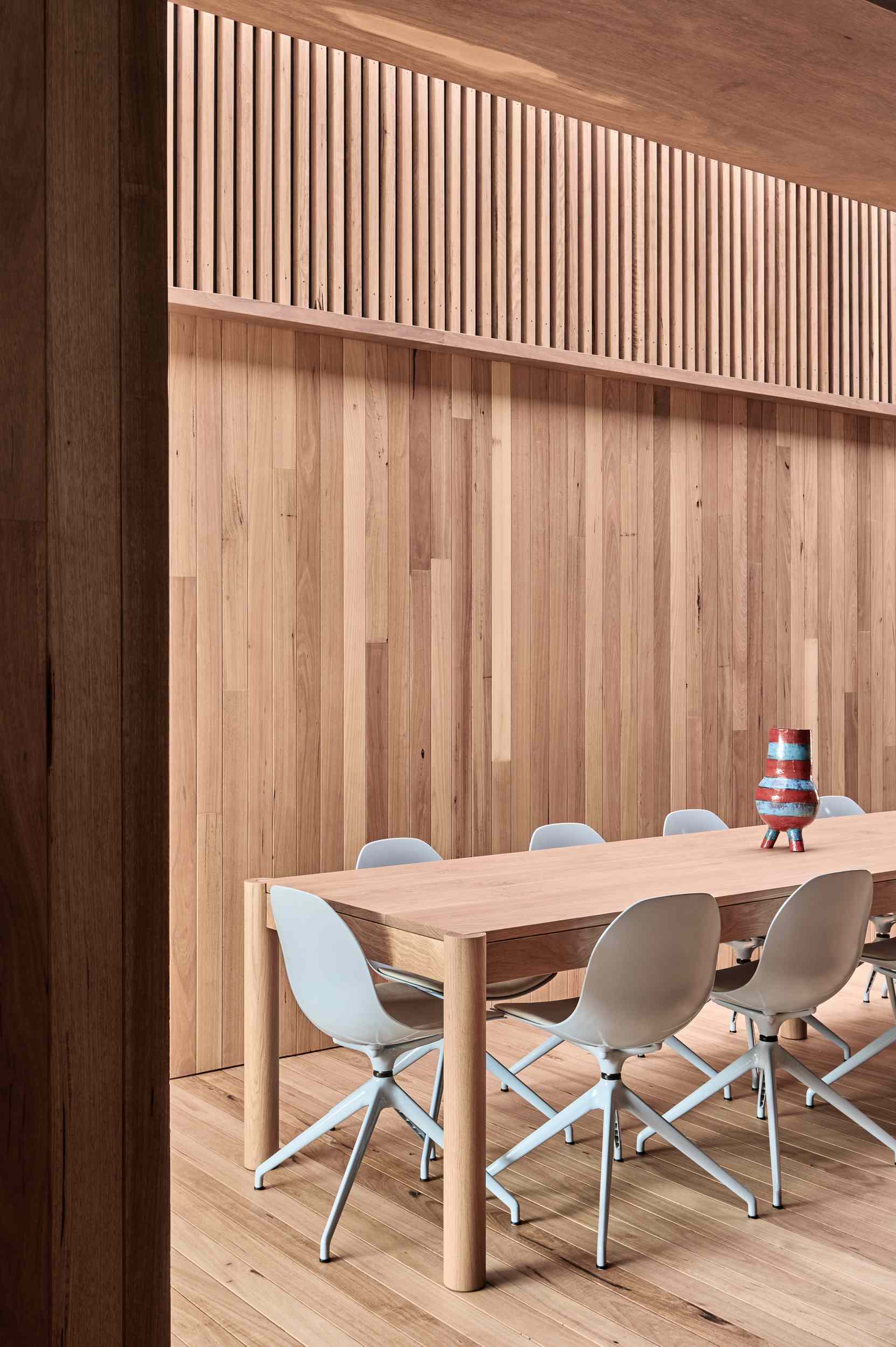 The interior boardwalk flows straight into the dining area, where timber walls and flooring create a warm, cohesive feel. A long wooden table anchors the space, paired with soft grey chairs that add a gentle contrast to the rich natural tones.