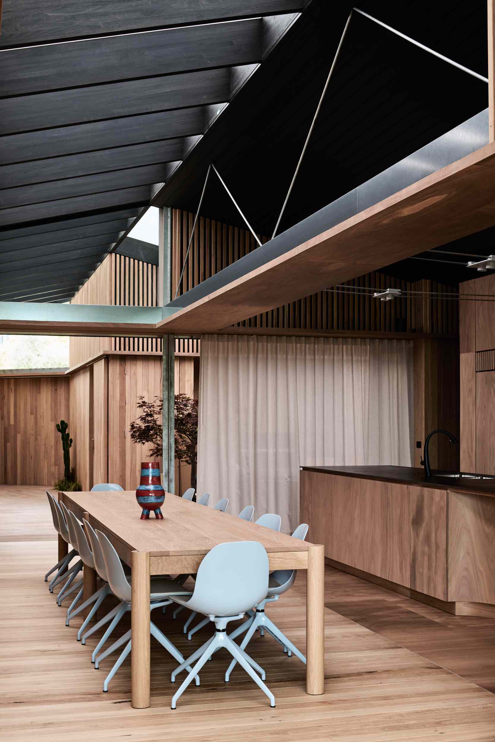 The interior boardwalk flows straight into the dining area, where timber walls and flooring create a warm, cohesive feel. A long wooden table anchors the space, paired with soft grey chairs that add a gentle contrast to the rich natural tones.